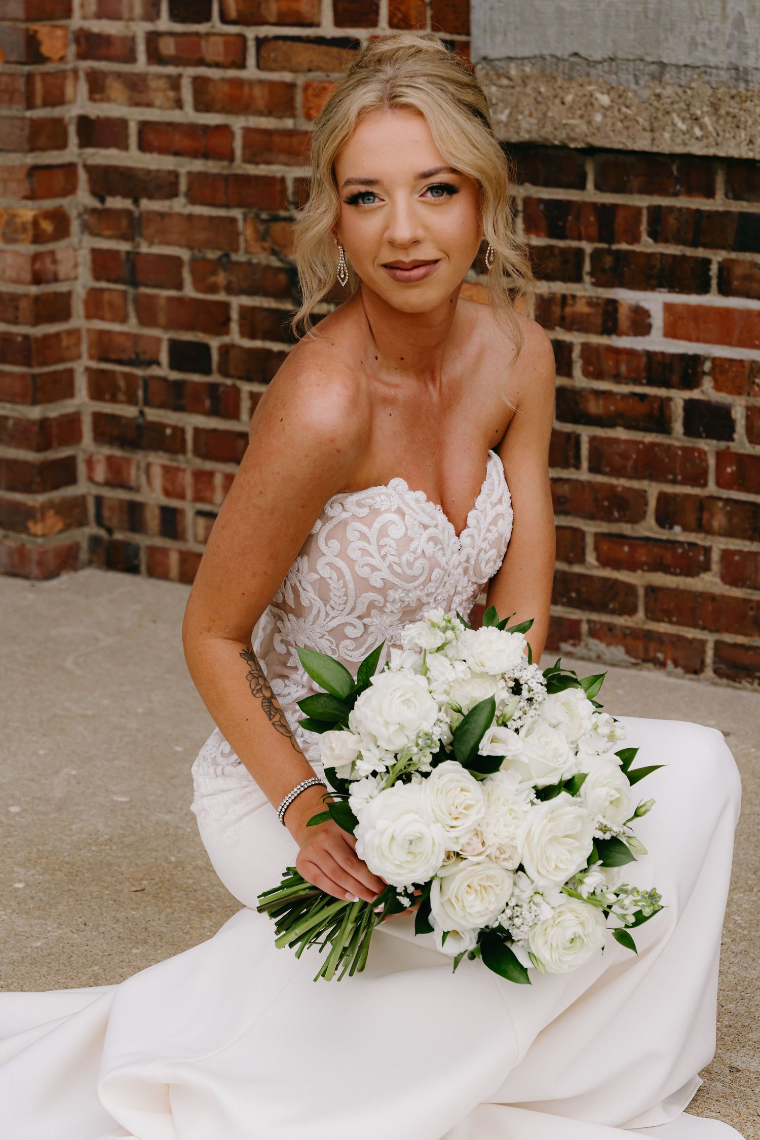 Bride in lace dress holds elegant white floral bouquet against brick background.