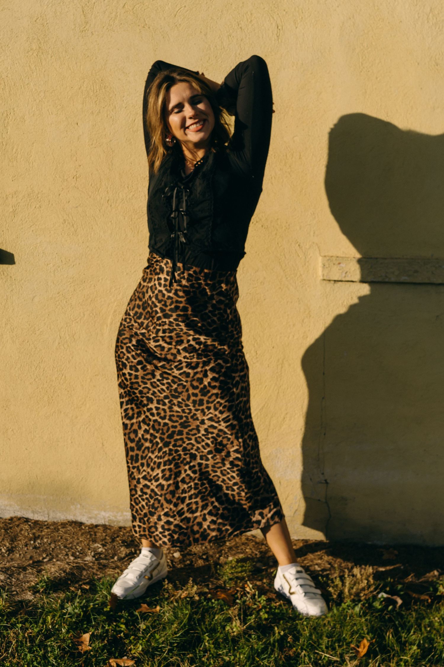 Person in leopard print skirt and black top poses playfully against beige wall with white sneakers.
