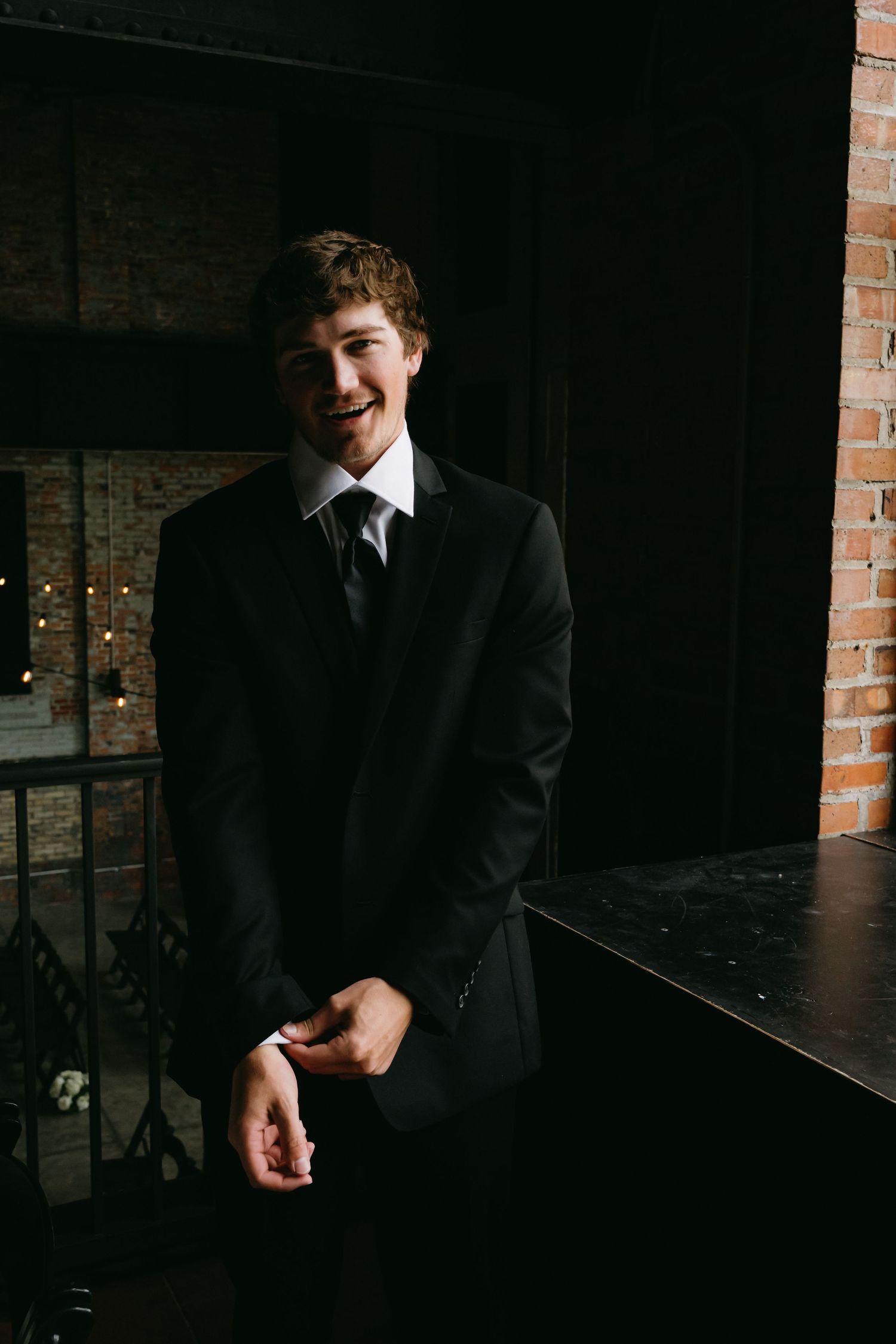 Groom in black suit adjusts his tie in a dimly lit room with brick walls.