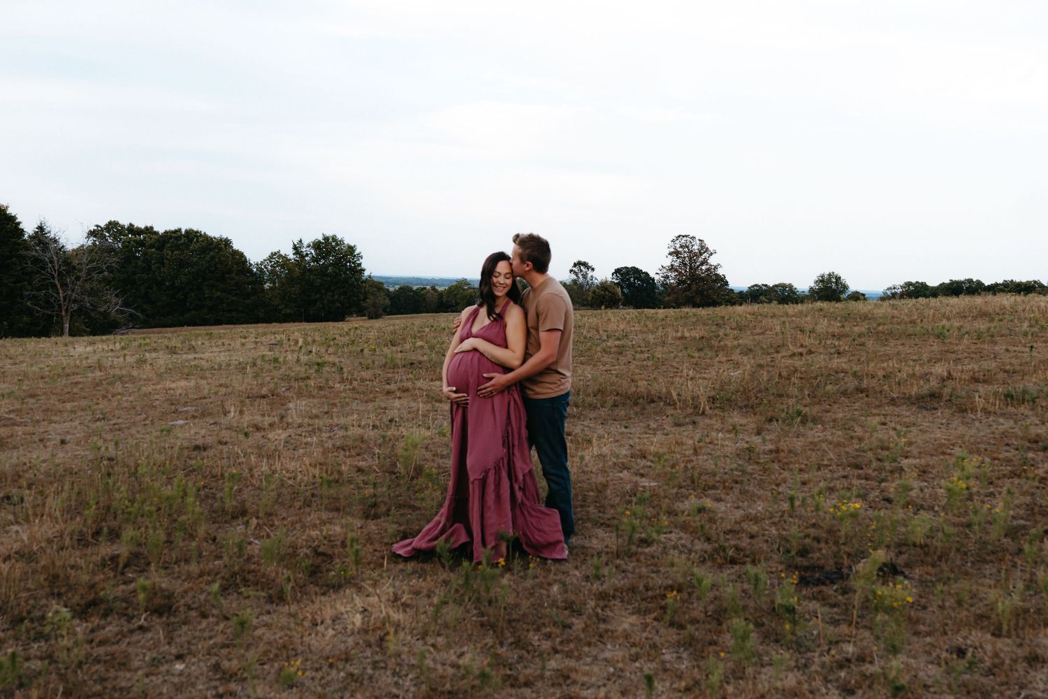 A couple in burgundy attire embraces in a rustic field during golden hour.