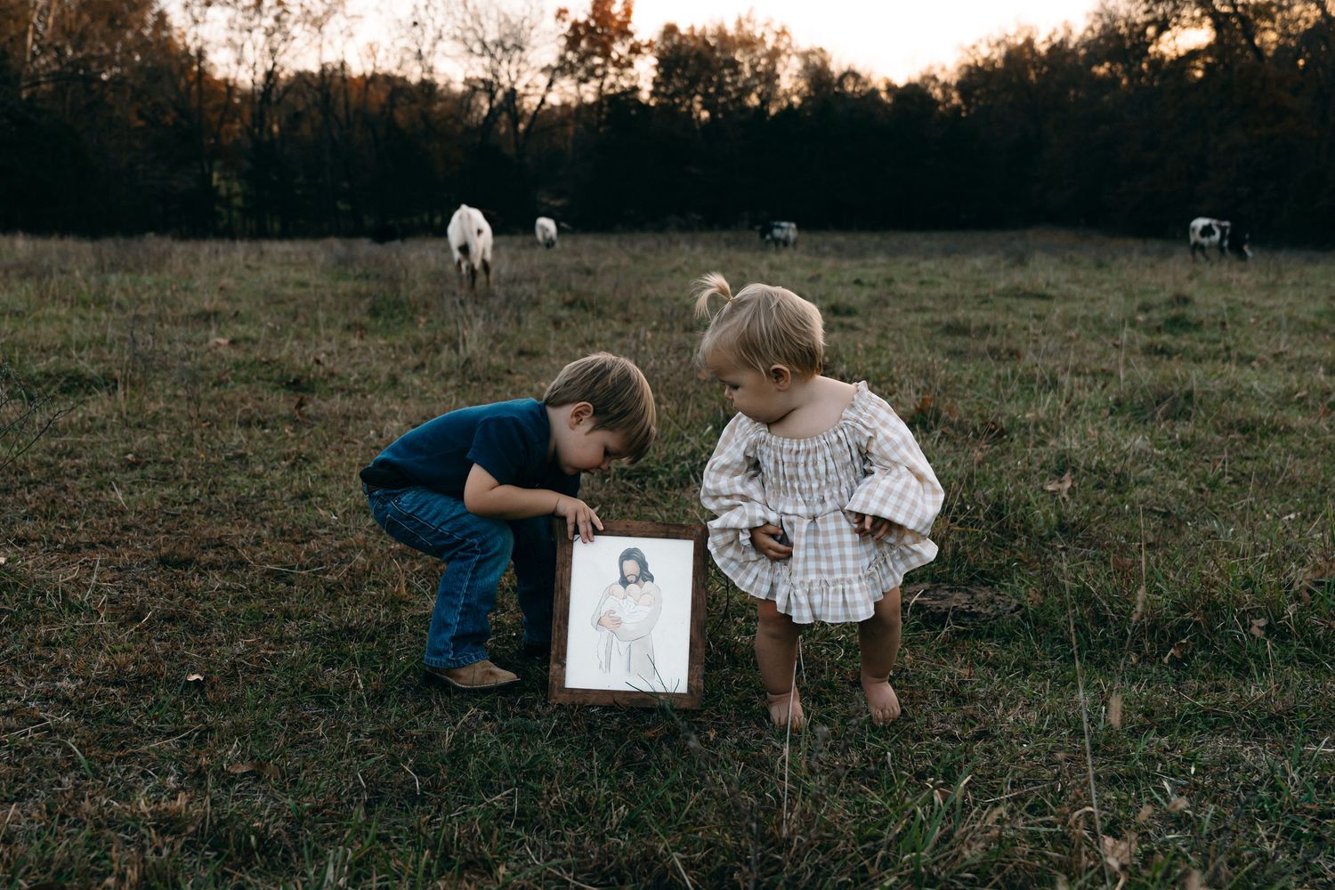 Two children examine a picture frame while sitting in a grassy field with animals in the background.