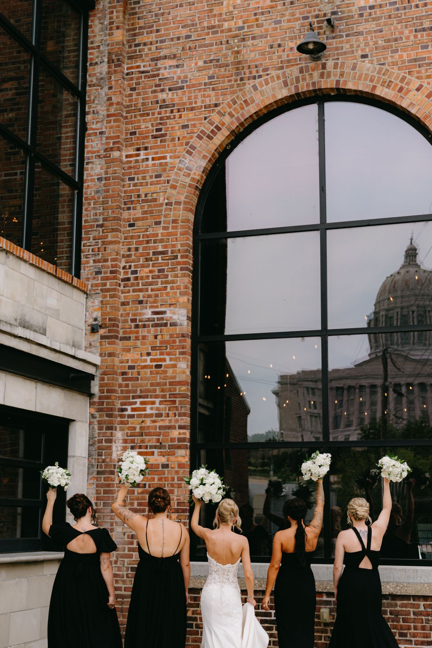 Wedding party in black dresses stand against brick building with dome visible through windows.