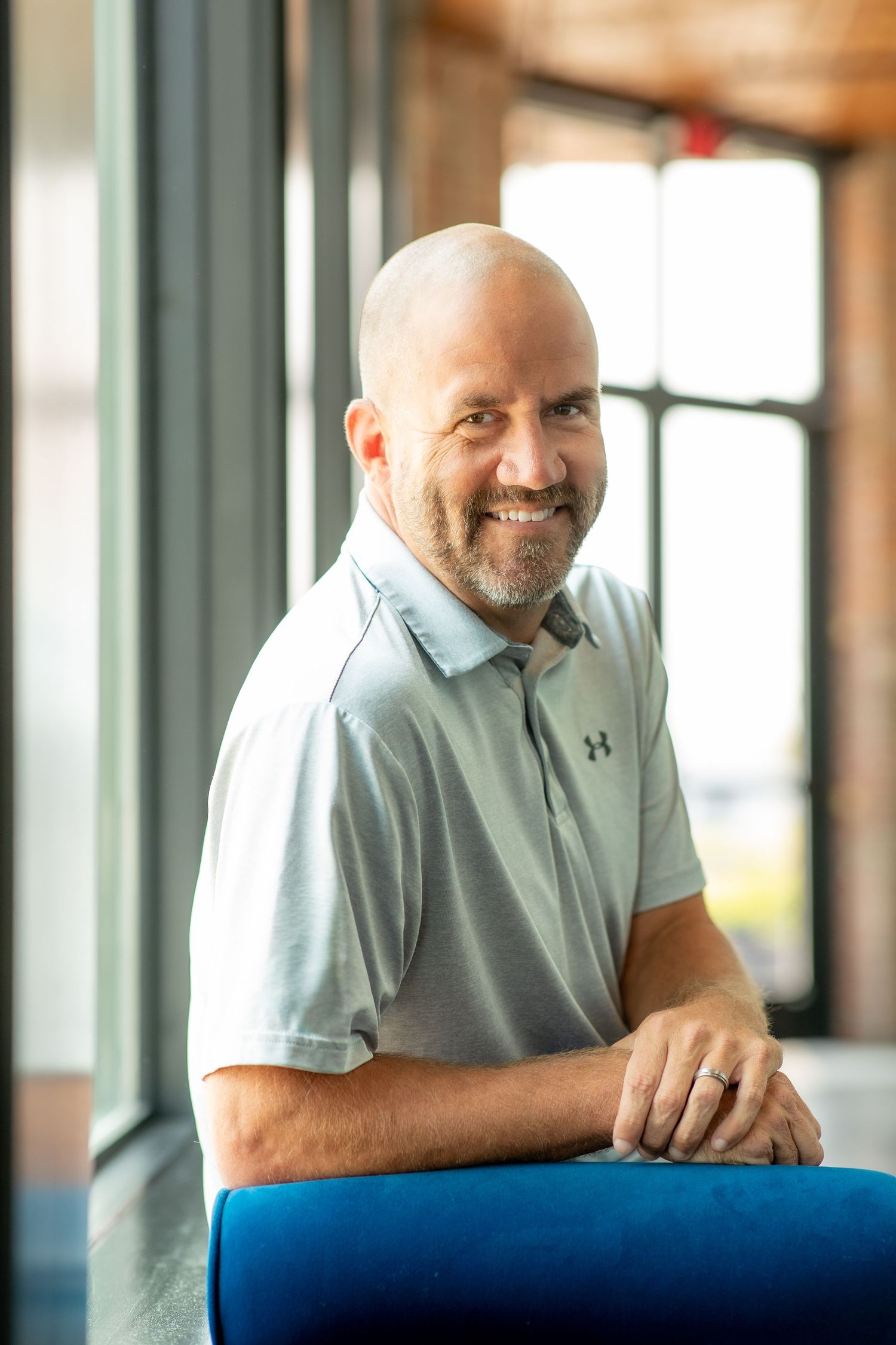 Man in light blue polo shirt smiles while leaning on blue chair in bright indoor setting.