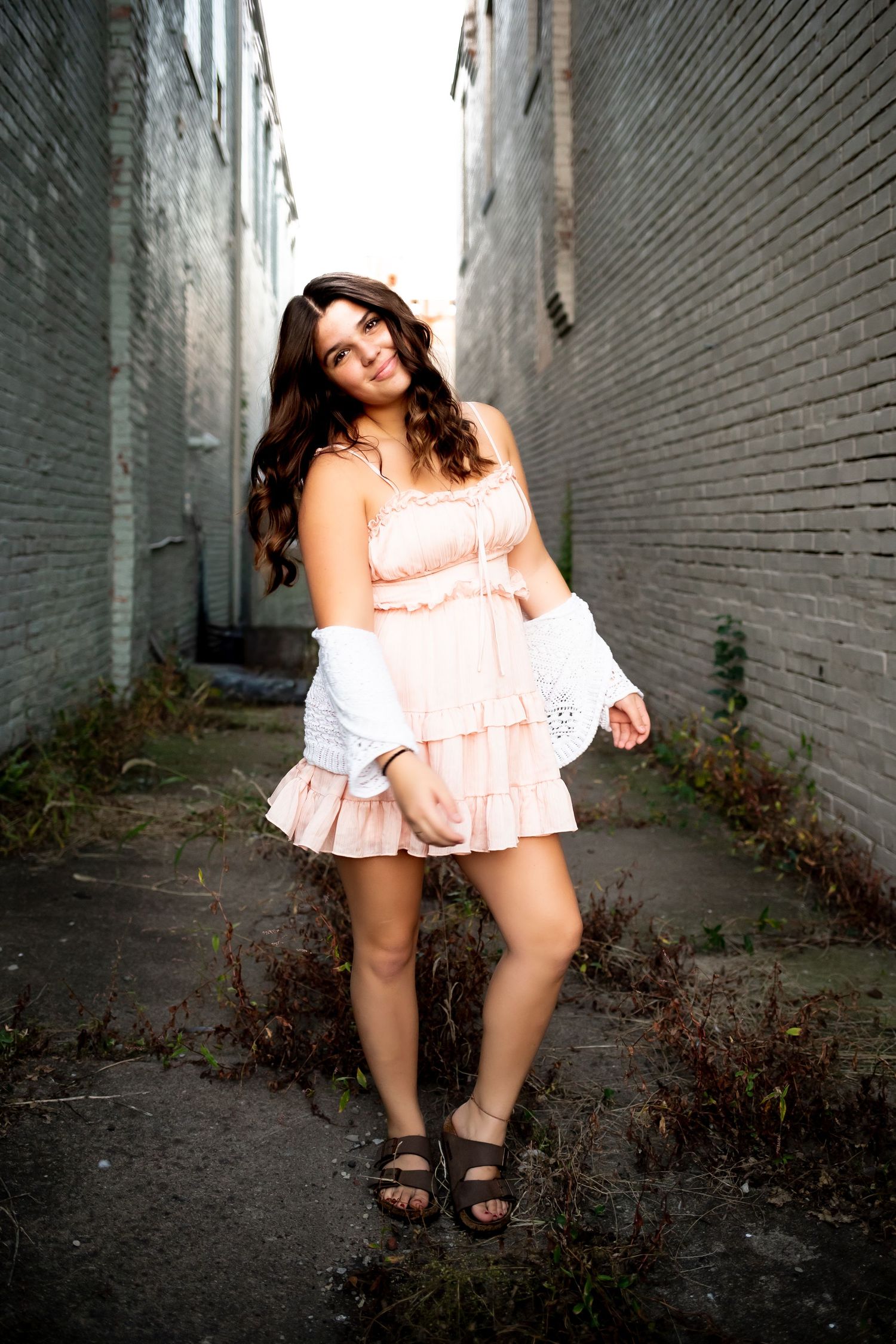 A person in a pink ruffled dress poses against brick walls in an urban alleyway setting.