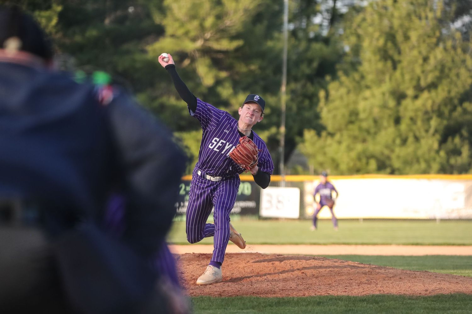 Baseball pitcher in purple uniform follows through on delivery from pitcher's mound during game action.