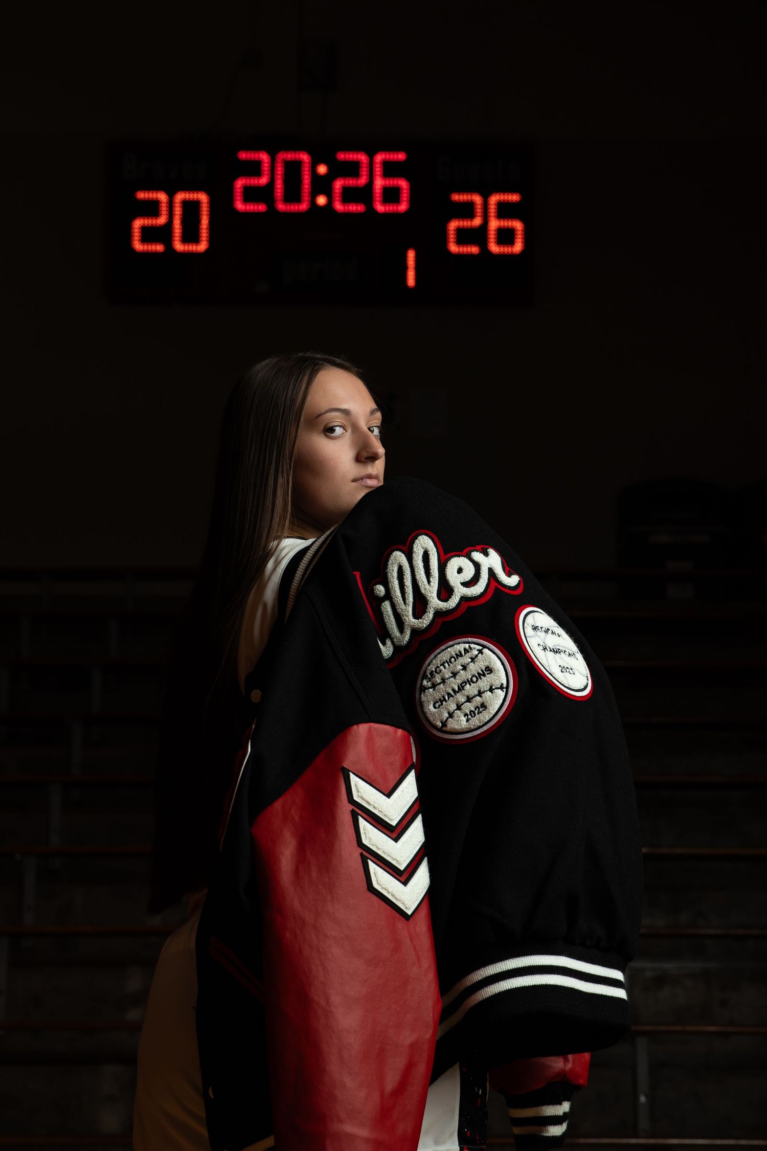 A person in a black and red varsity jacket poses against a dark background with a digital clock display.