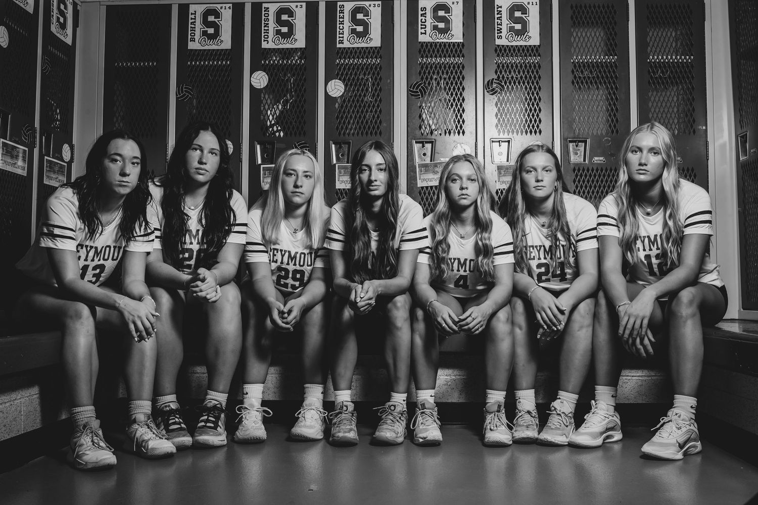 High school volleyball team players sit together on locker room bench in black and white photo wearing matching uniforms.