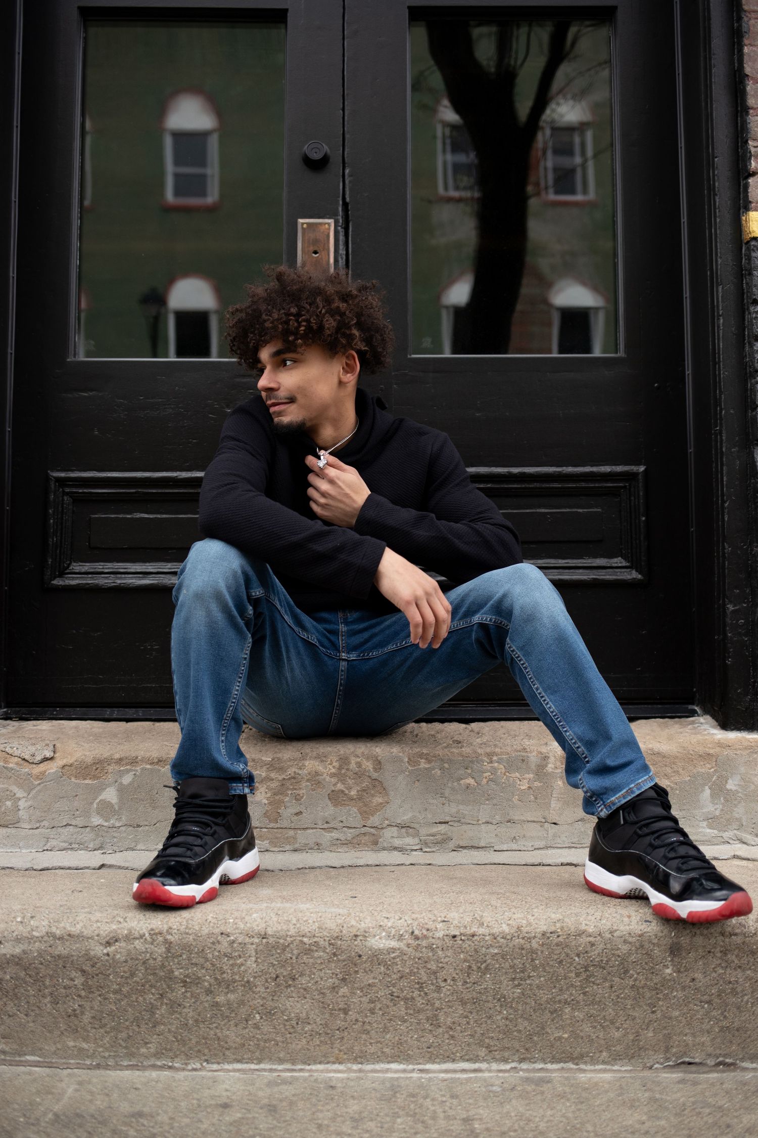 Person in black top and jeans sits casually on concrete curb wearing red and black sneakers.