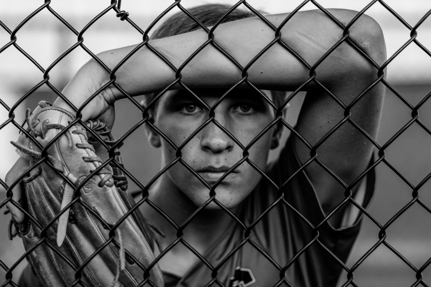 A black and white portrait through a chain link fence shows a figure wearing a hat with an intense gaze.