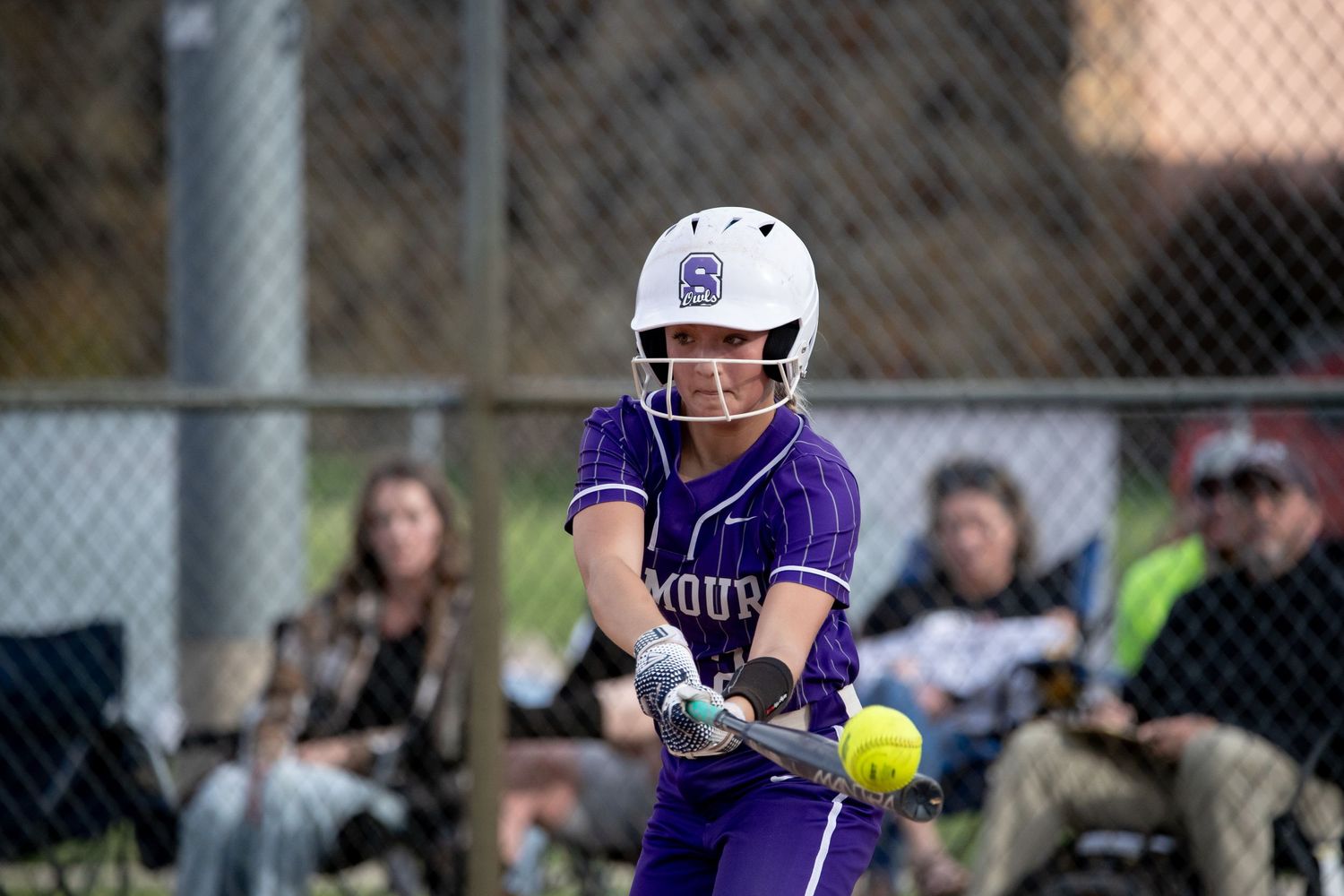 Softball player in purple uniform hits yellow ball during competitive game with chain link fence in background.