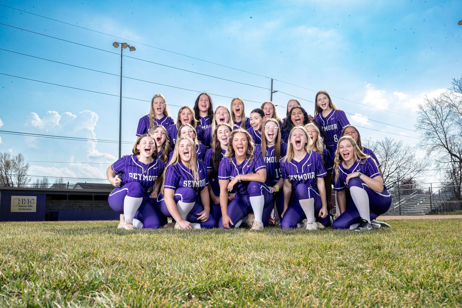 A group wearing matching purple jerseys poses together on a grassy sports field on a sunny day.