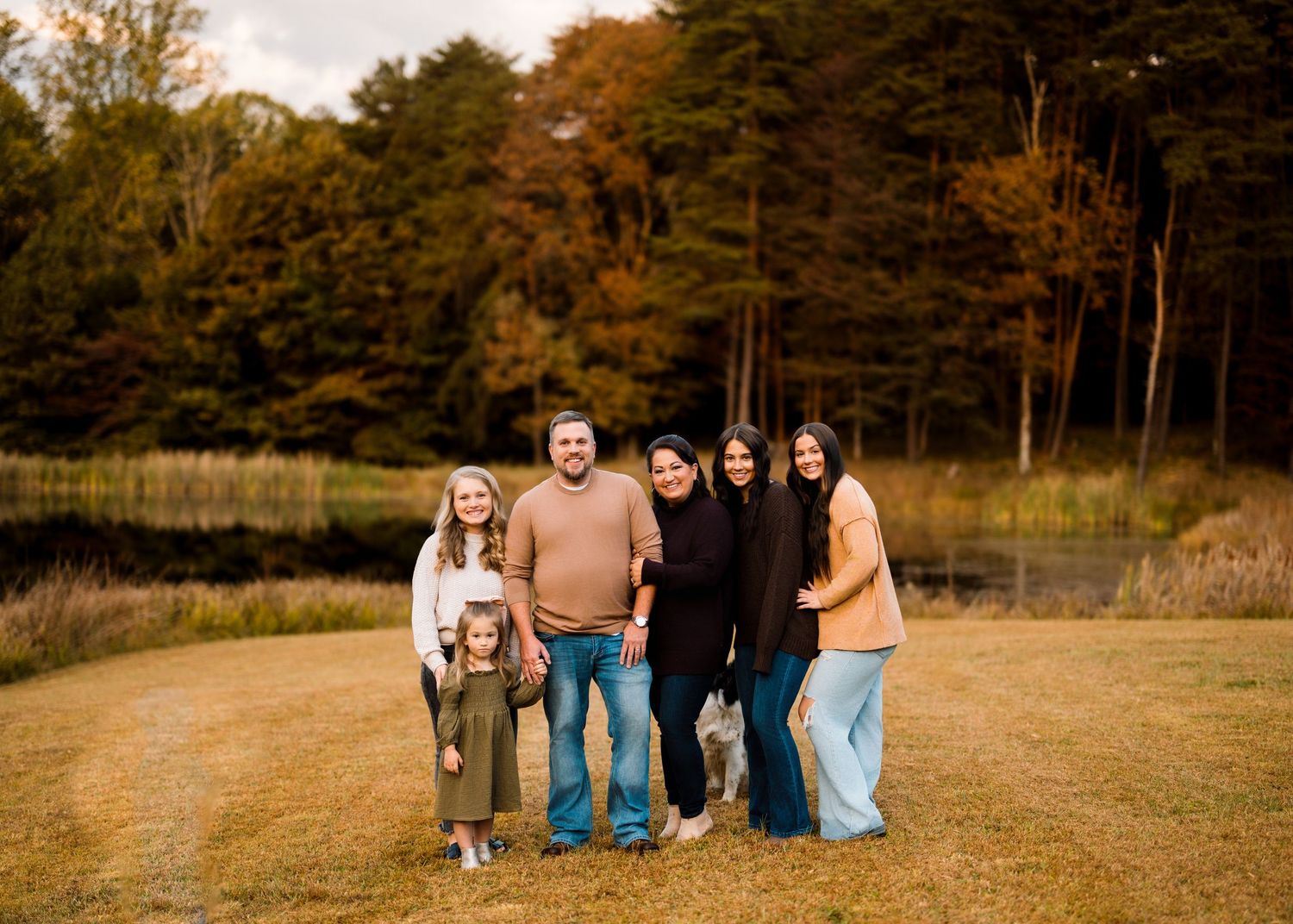 A group sits together in a golden autumn field with tall pine trees in the background during sunset.