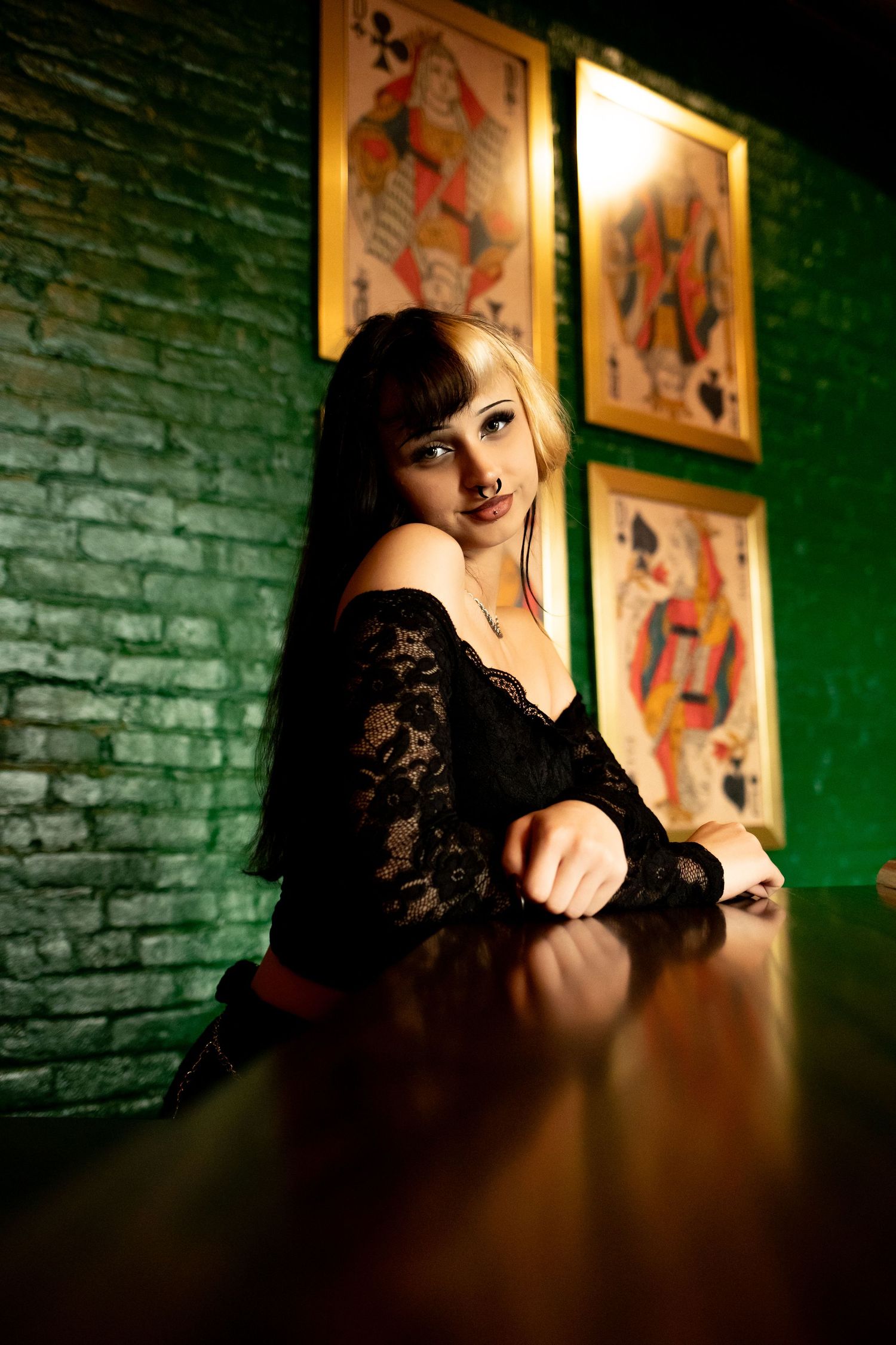 A woman in black lace sits at a bar with vintage art posters on the green brick wall behind her.