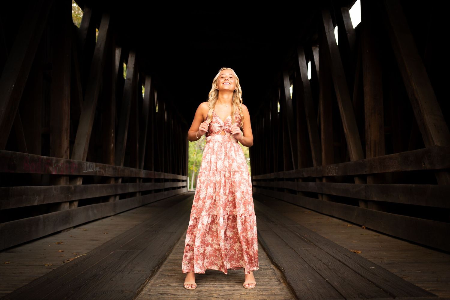 A person in a pink floral dress poses on a wooden bridge walkway in a romantic outdoor setting.