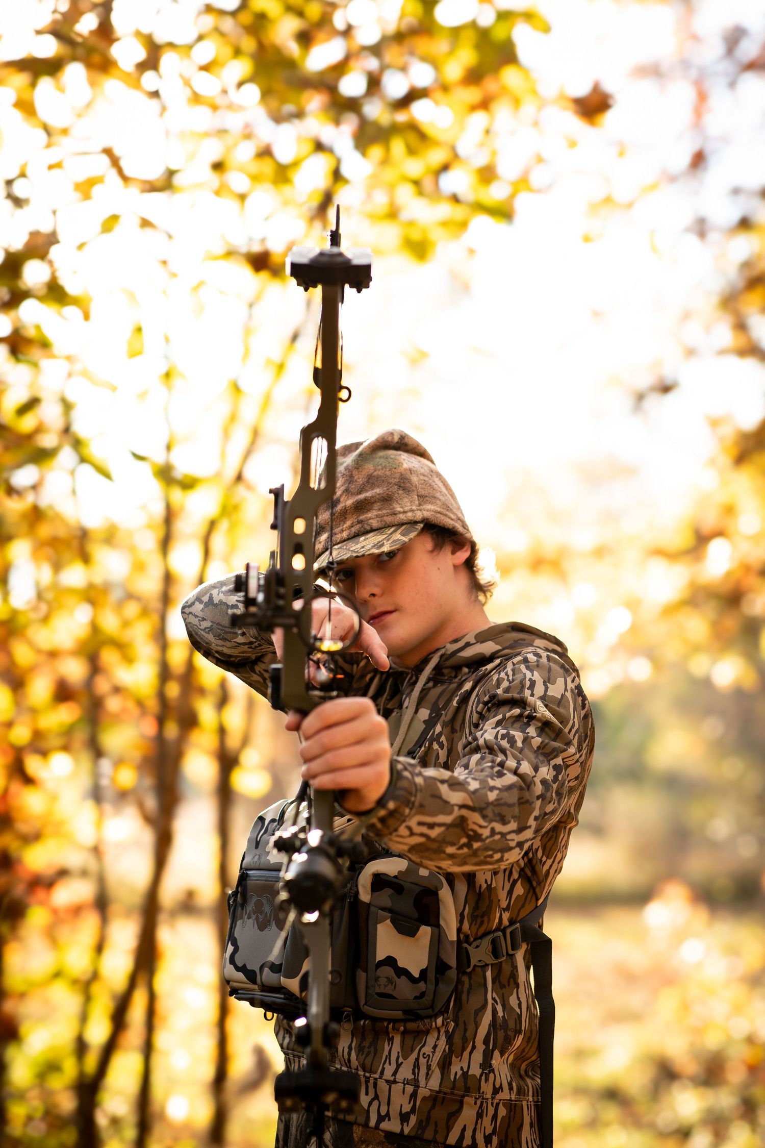 A hunter in camouflage gear holds a compound bow while standing in autumn woods with golden foliage.