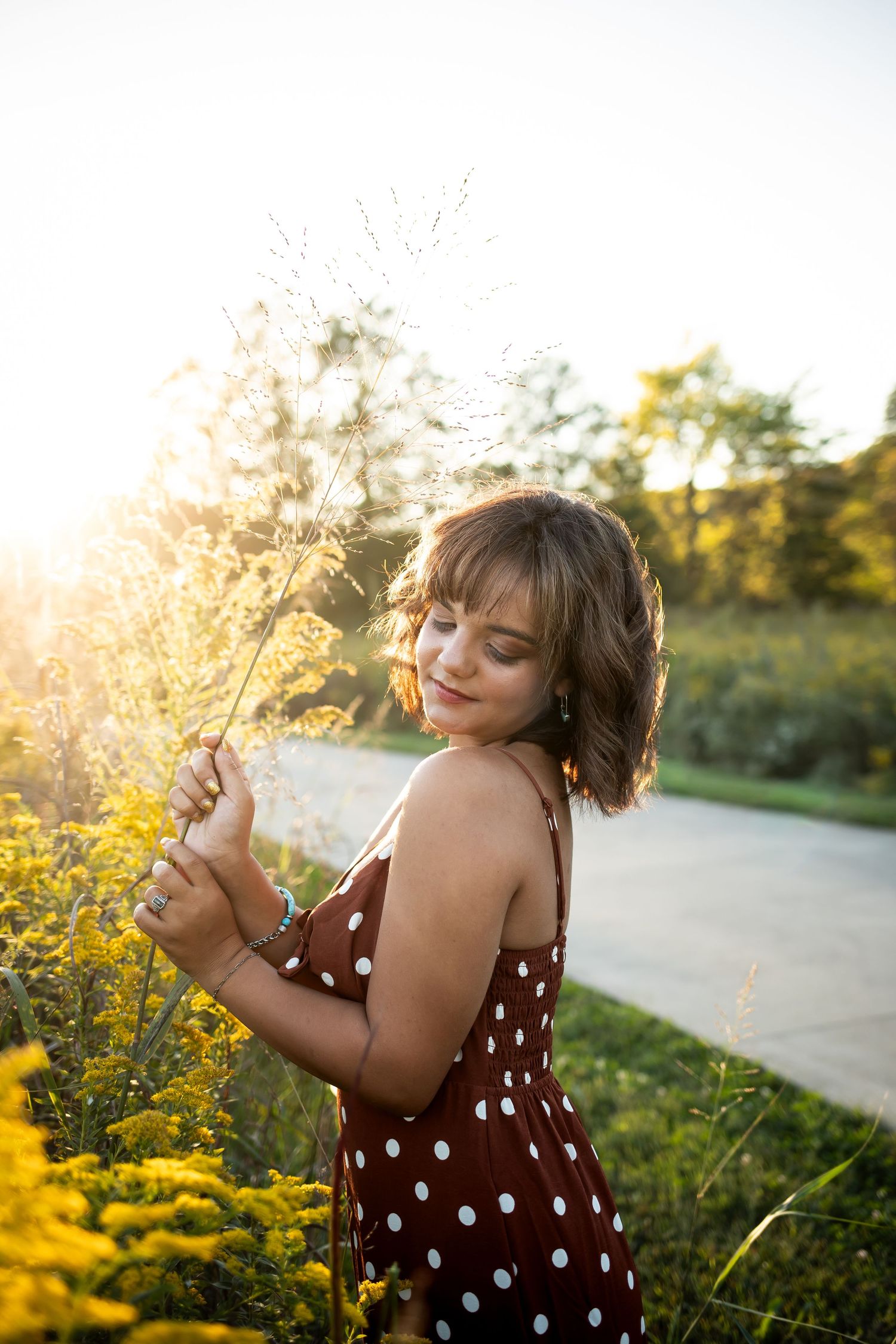 A person in a polka dot dress stands among yellow wildflowers at sunset holding dried plants on a summer evening.