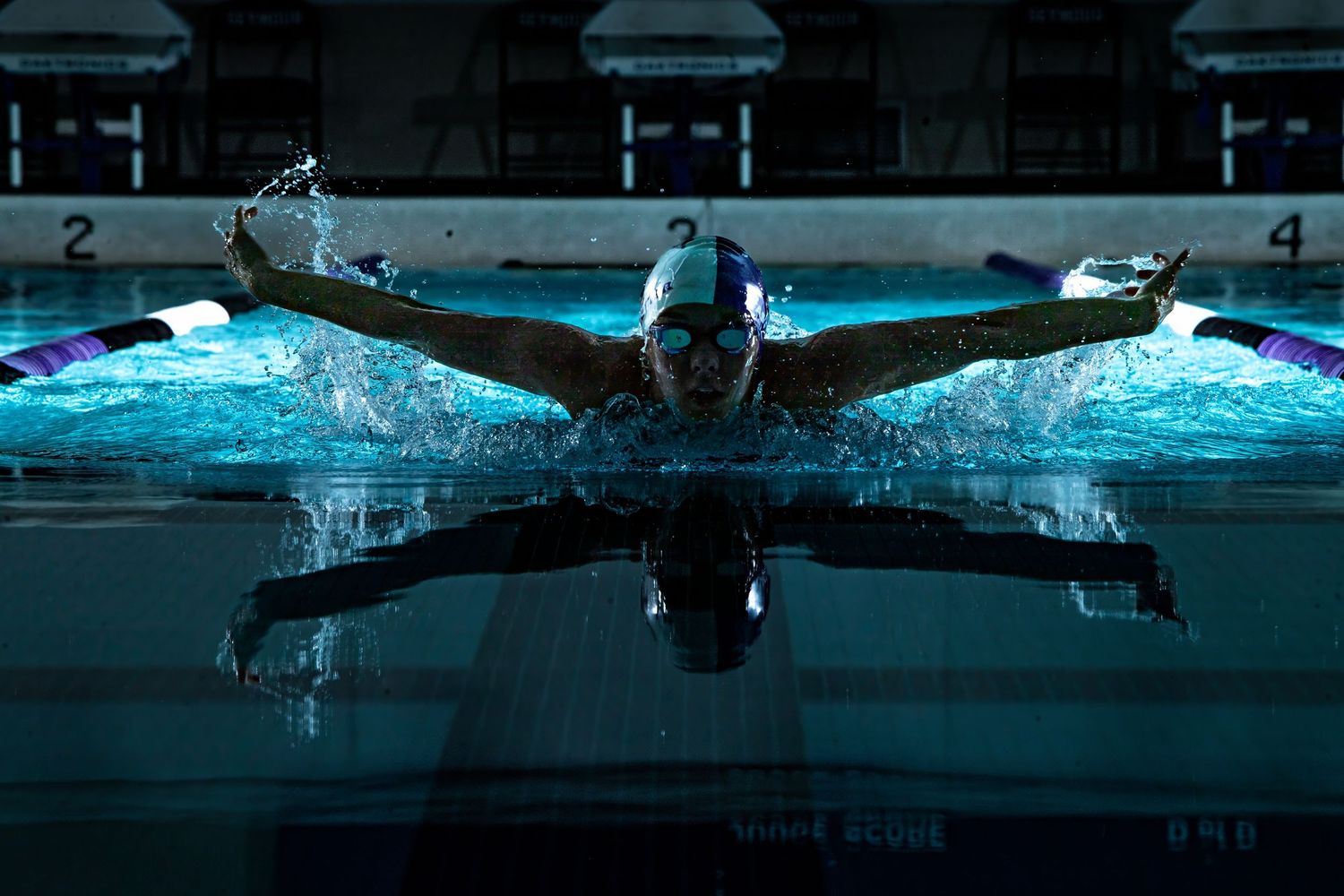 A swimmer performs the butterfly stroke in a pool with dramatic blue lighting creating a mirror-like reflection.