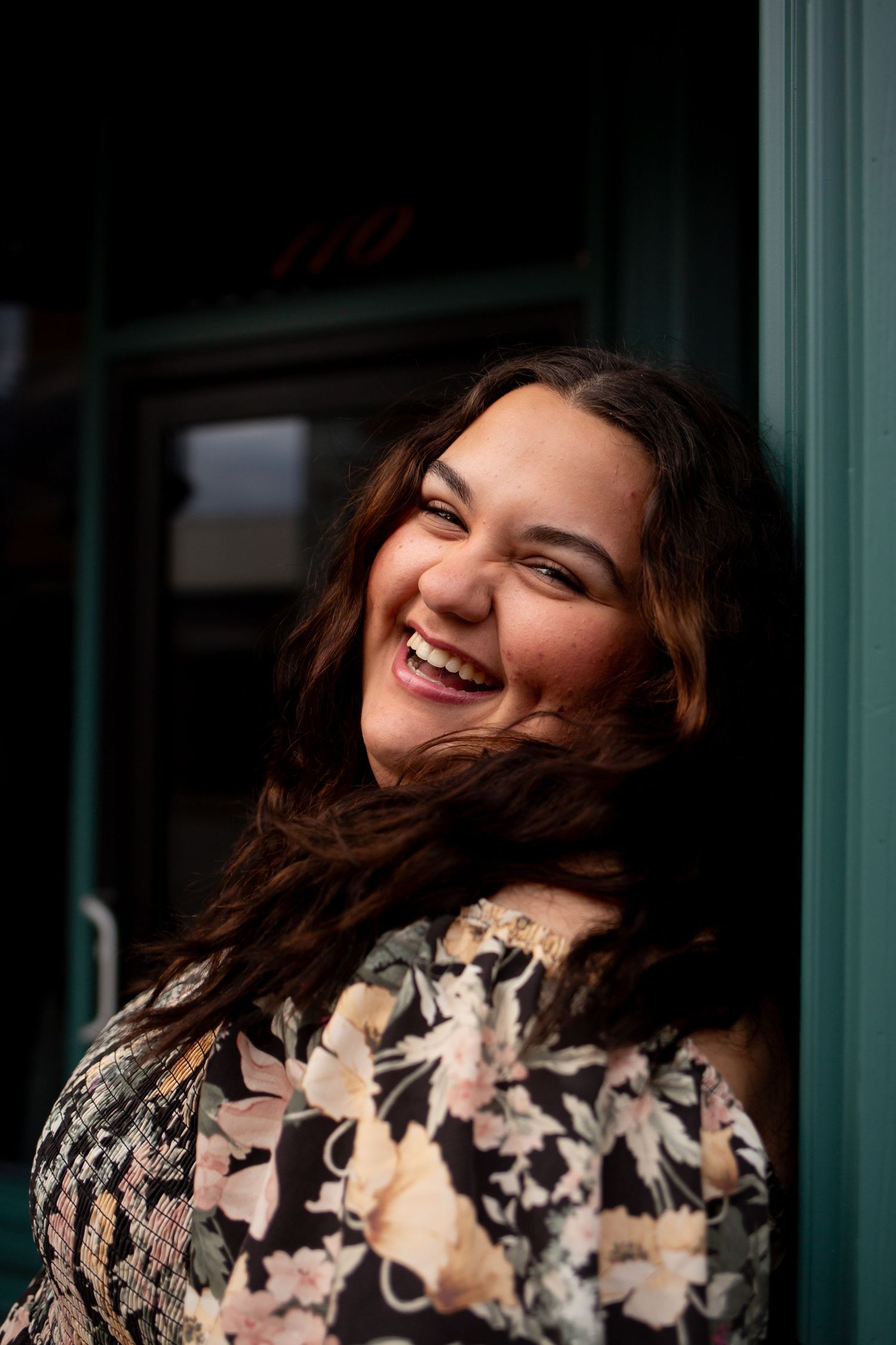 A joyful portrait against a teal background shows a genuine laugh and flowing brown hair in a floral print dress.