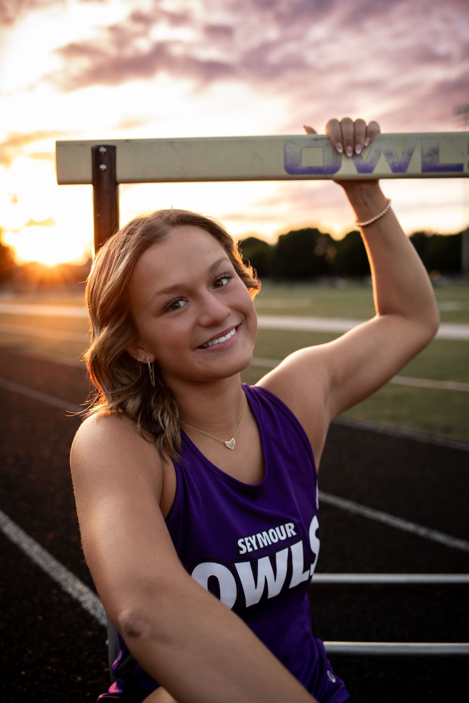 Athletic figure in purple jersey poses against sunset on sports field with dramatic sky in background.