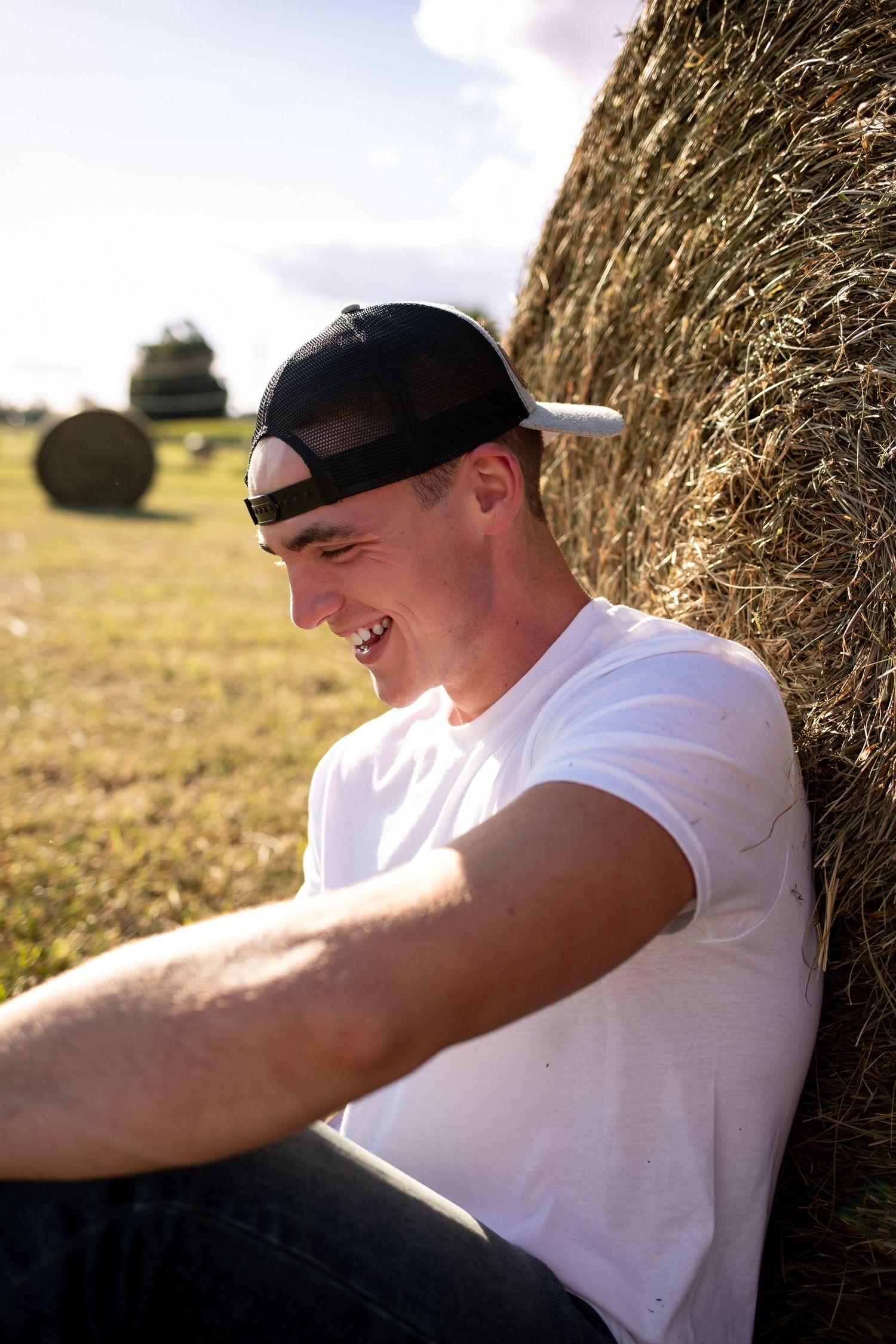 Person in white t-shirt relaxing against hay bale in sunny field with round bales visible in background.