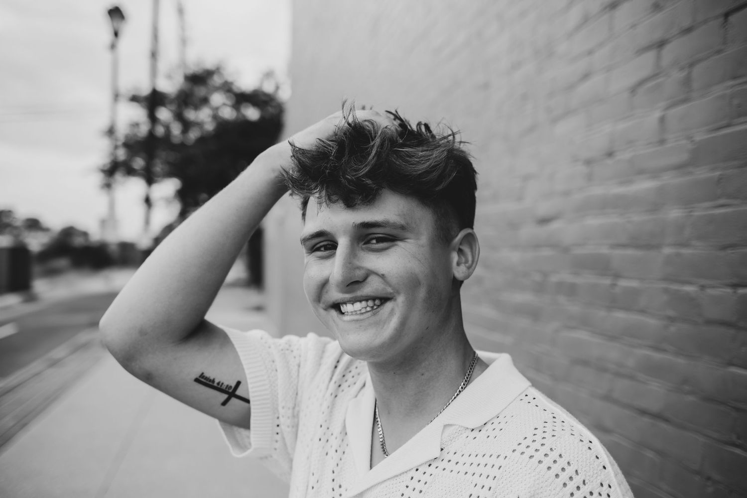 Black and white portrait of someone smiling while leaning against a white brick wall outdoors.