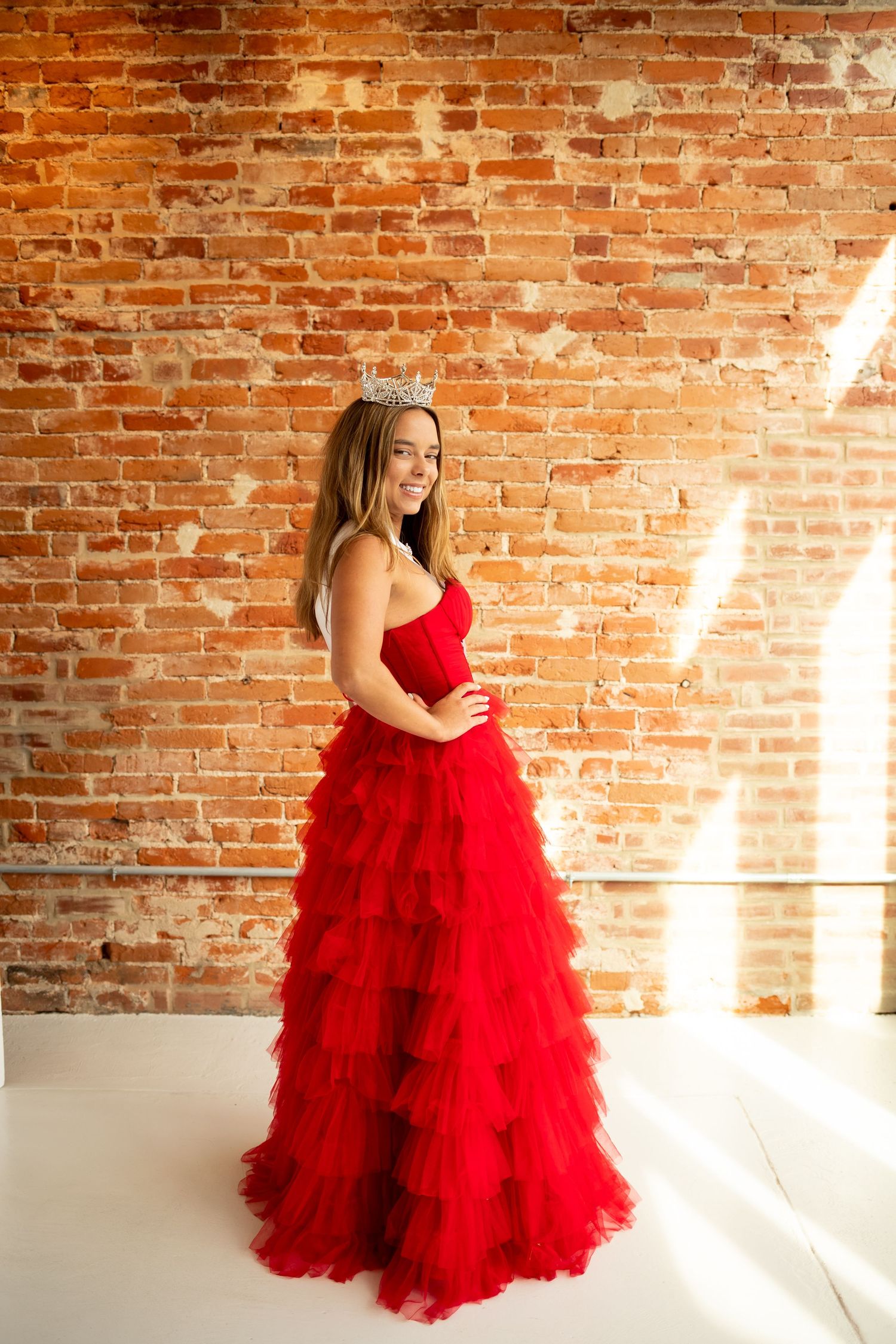 A stunning red tulle ballgown photographed in sequence against an exposed brick wall in a sunlit studio space.