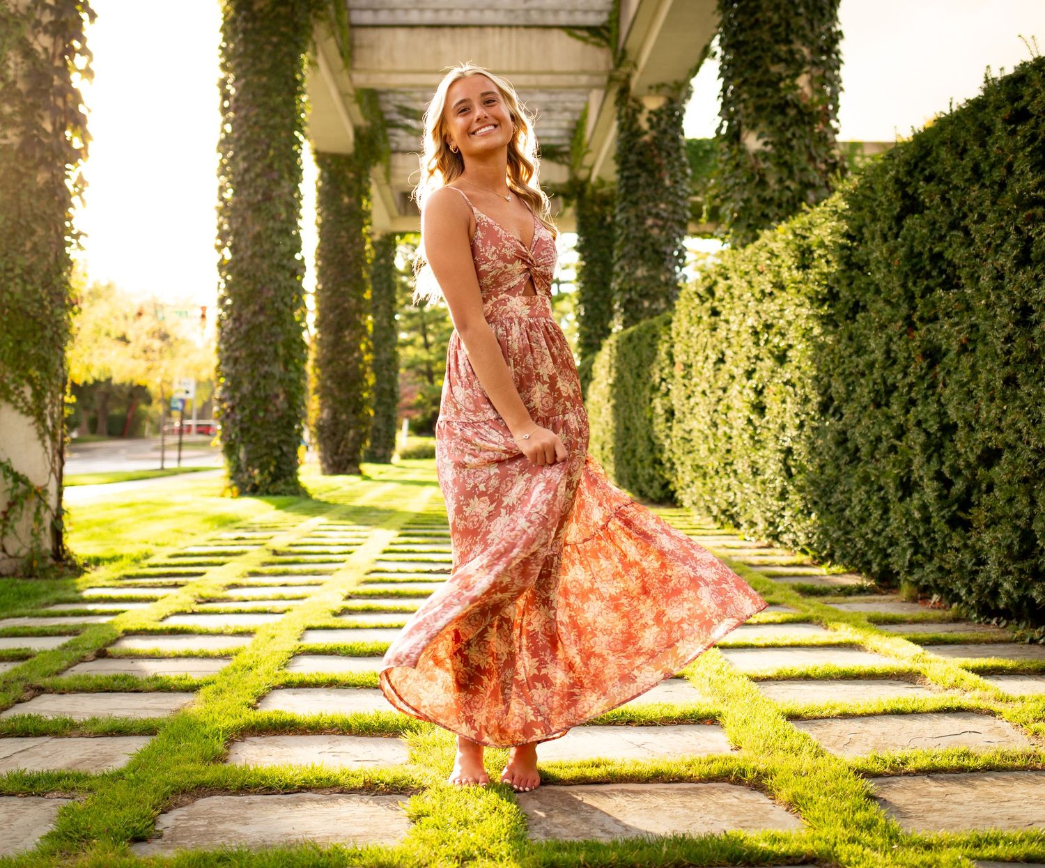 A person in a flowing floral dress twirls gracefully along a garden path lined with manicured hedges and stone pavers.