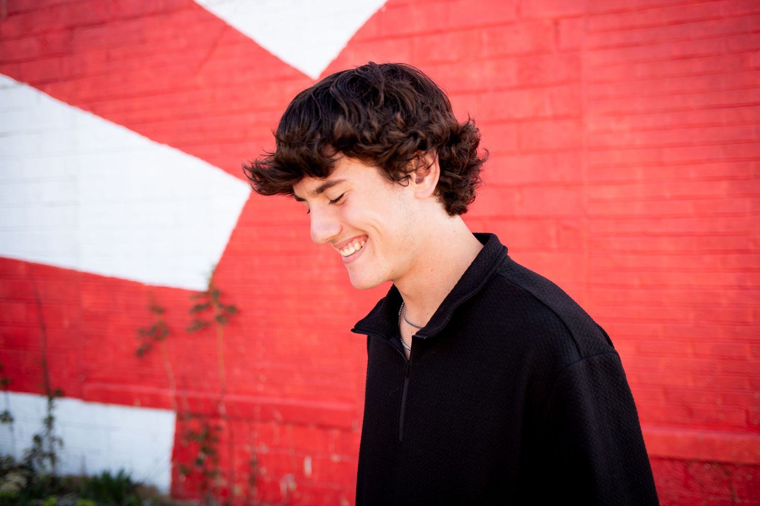 A person in a black jacket stands against a vibrant red brick wall in an outdoor setting.