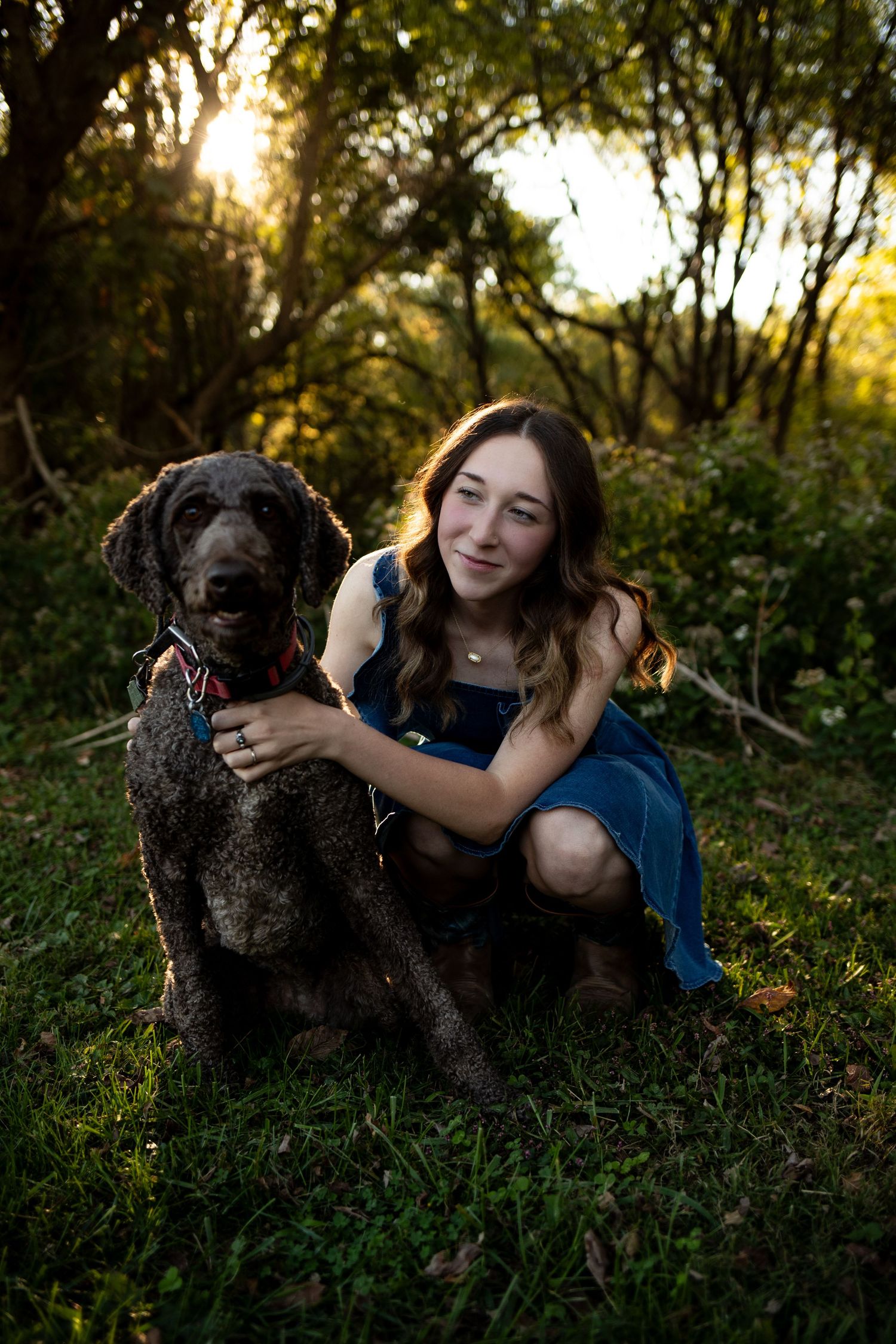 A person in a blue shirt lies in grass with a black poodle mix dog at sunset in a wooded area.