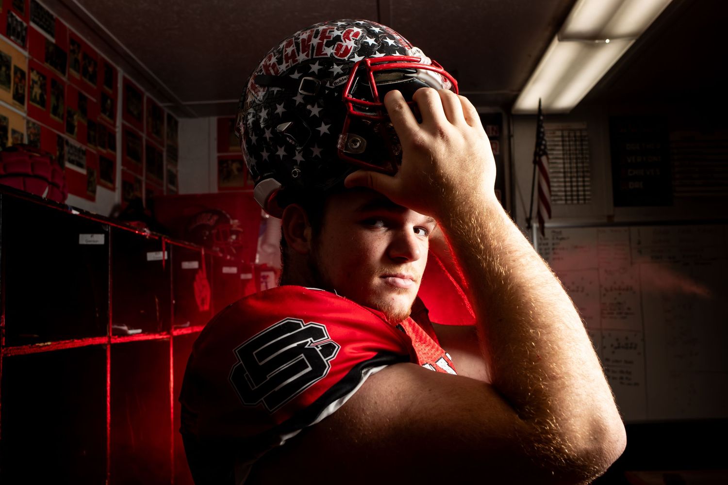 Football player adjusts black helmet with red accents in dark locker room with dramatic lighting.