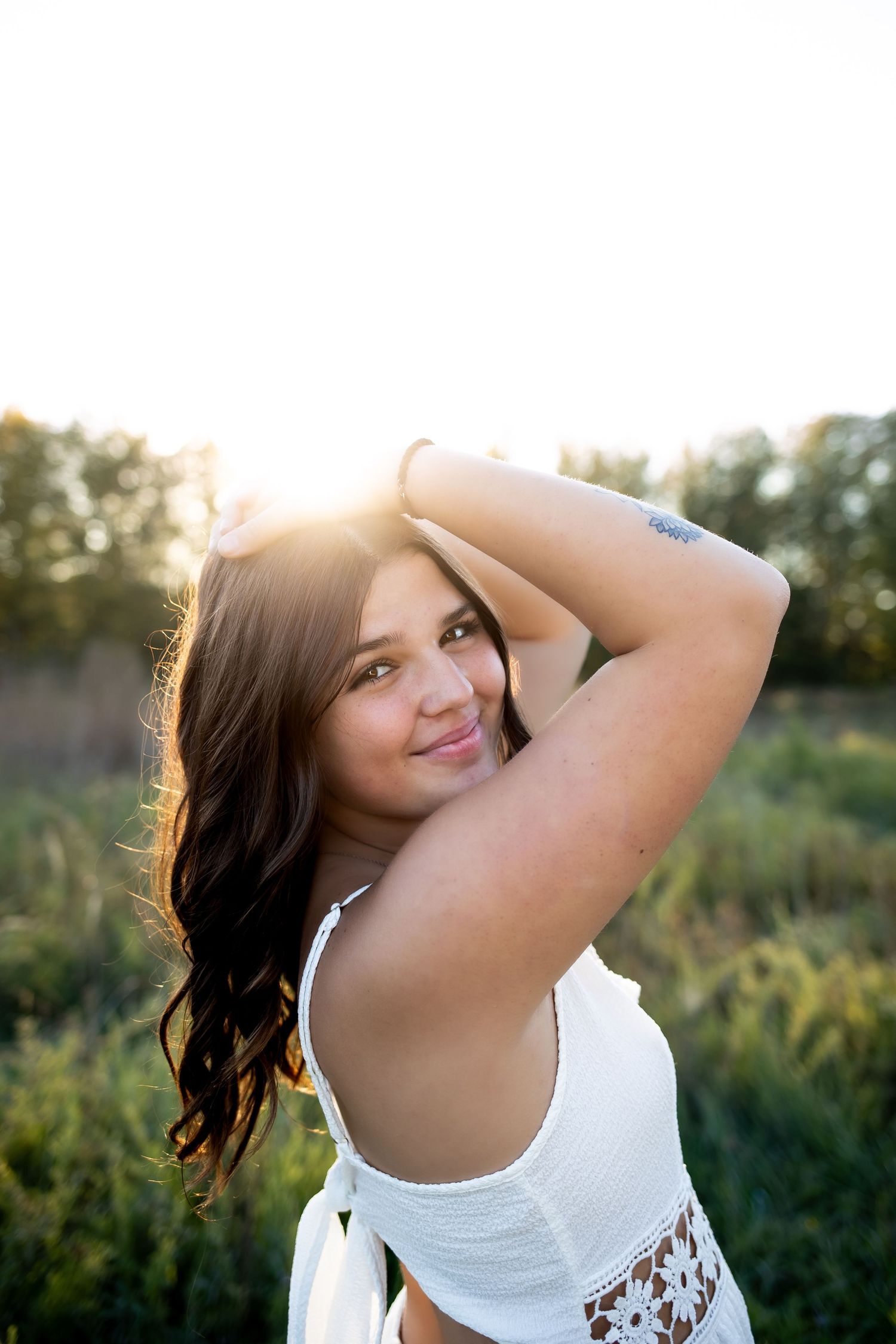 A woman in a white top poses outdoors at golden hour with sunlit foliage in the background.