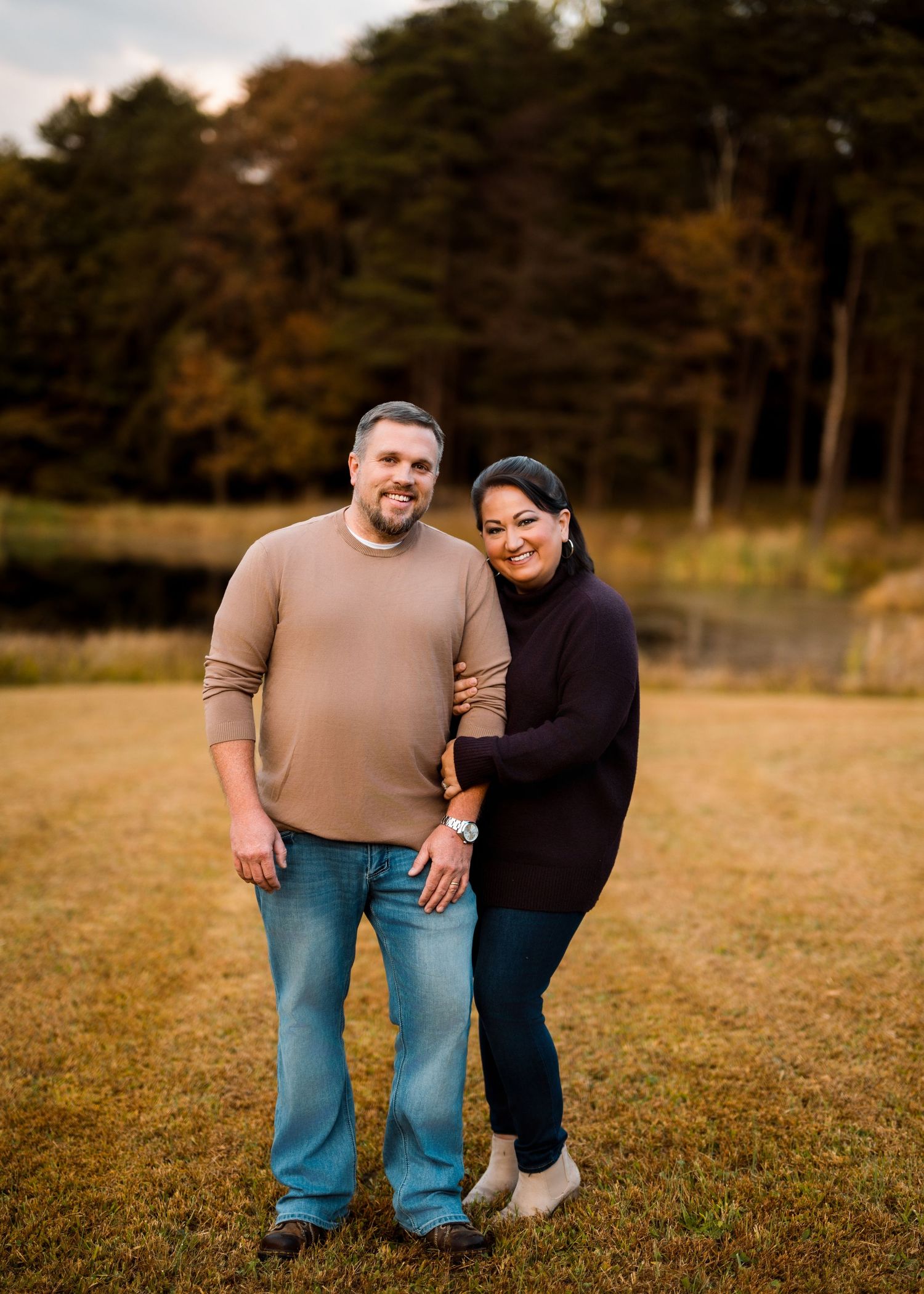 A couple stands together in a grassy field during autumn with pine trees in the background.