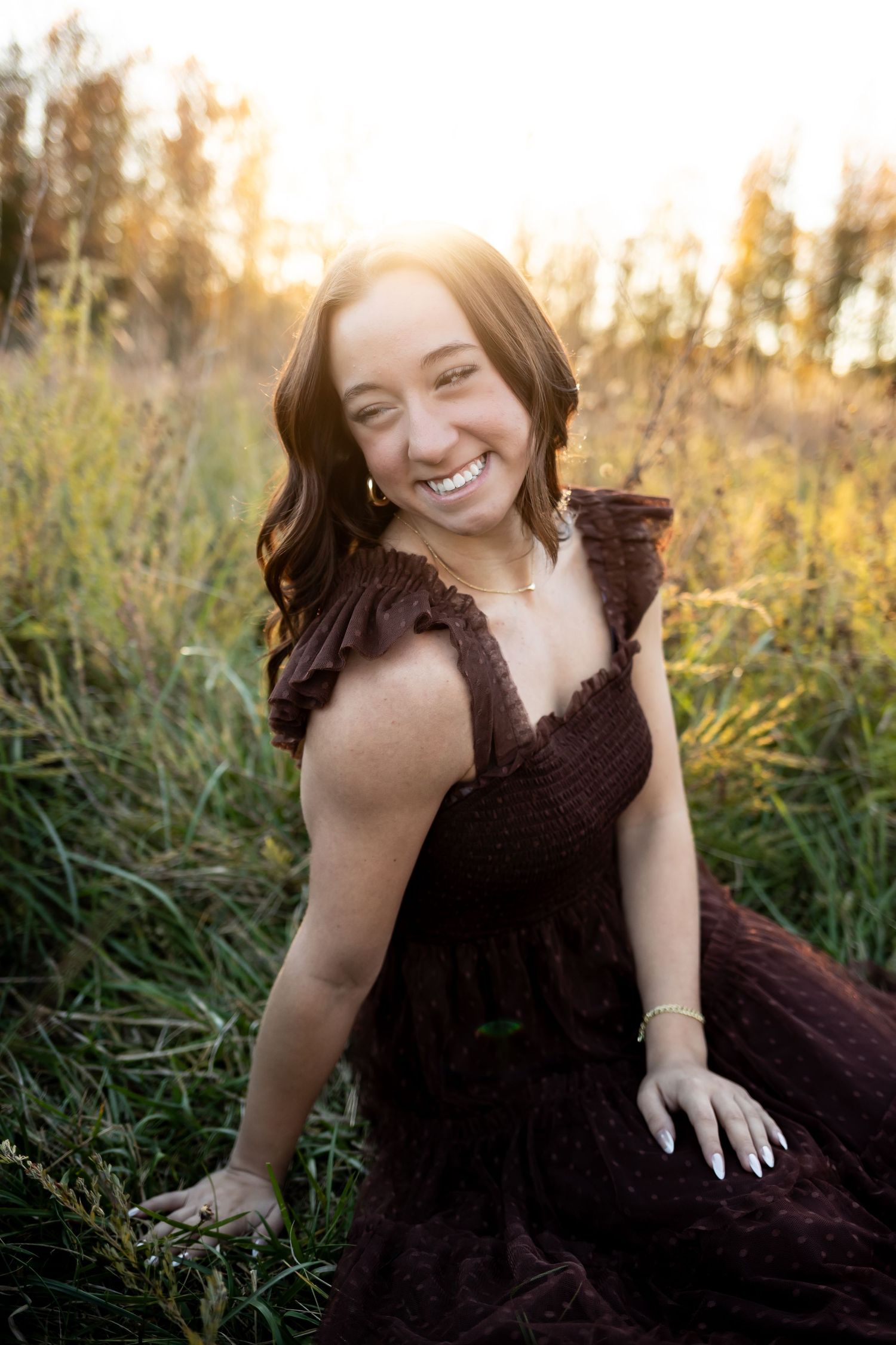 A joyful woman in a dark formal dress sits in tall grass during golden hour sunset lighting.