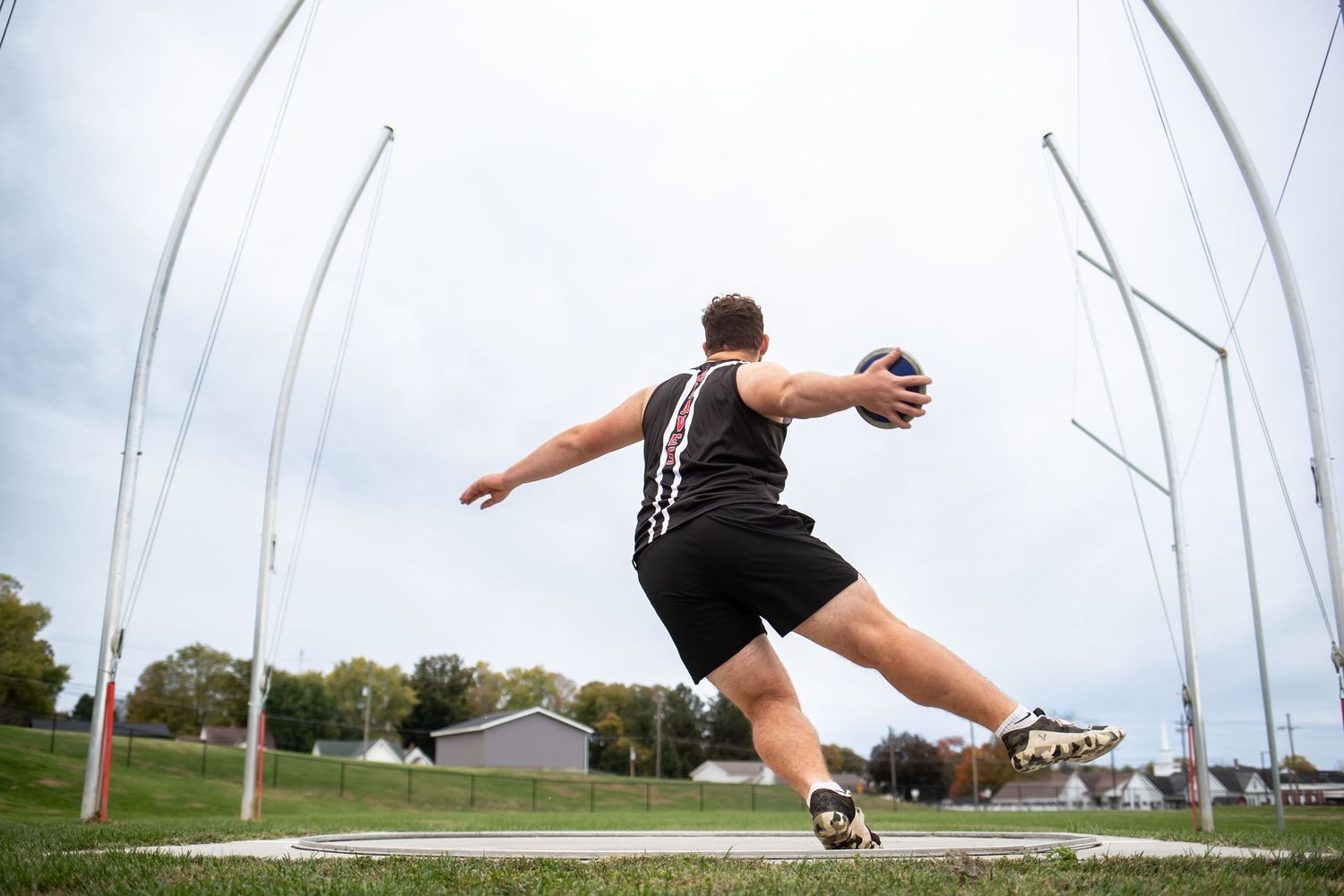 Track and field athlete in black uniform performs discus throw technique inside throwing cage at outdoor facility.