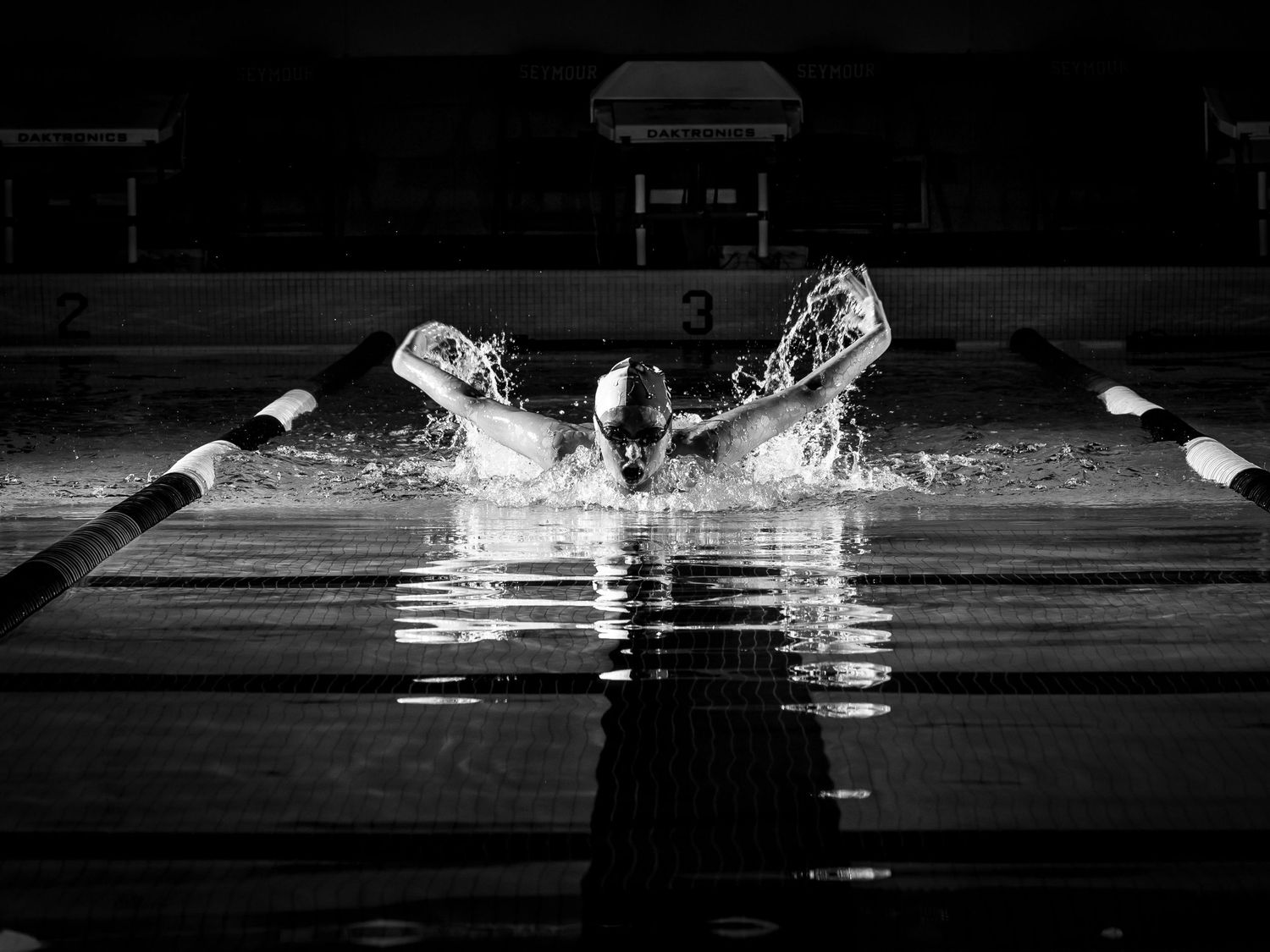 A dramatic black and white photograph captures a swimmer splashing through water in a pool with lane lines visible.