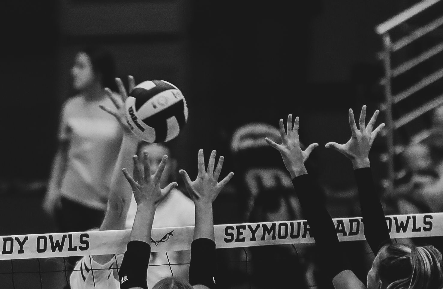 Black and white photo volleyball players raising their hands during a game with scoreboard visible in background.