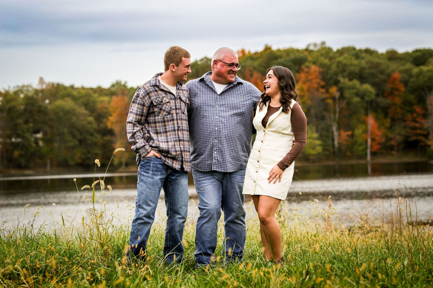 Three people stand together laughing in a grassy field with autumn trees and a lake in the background.