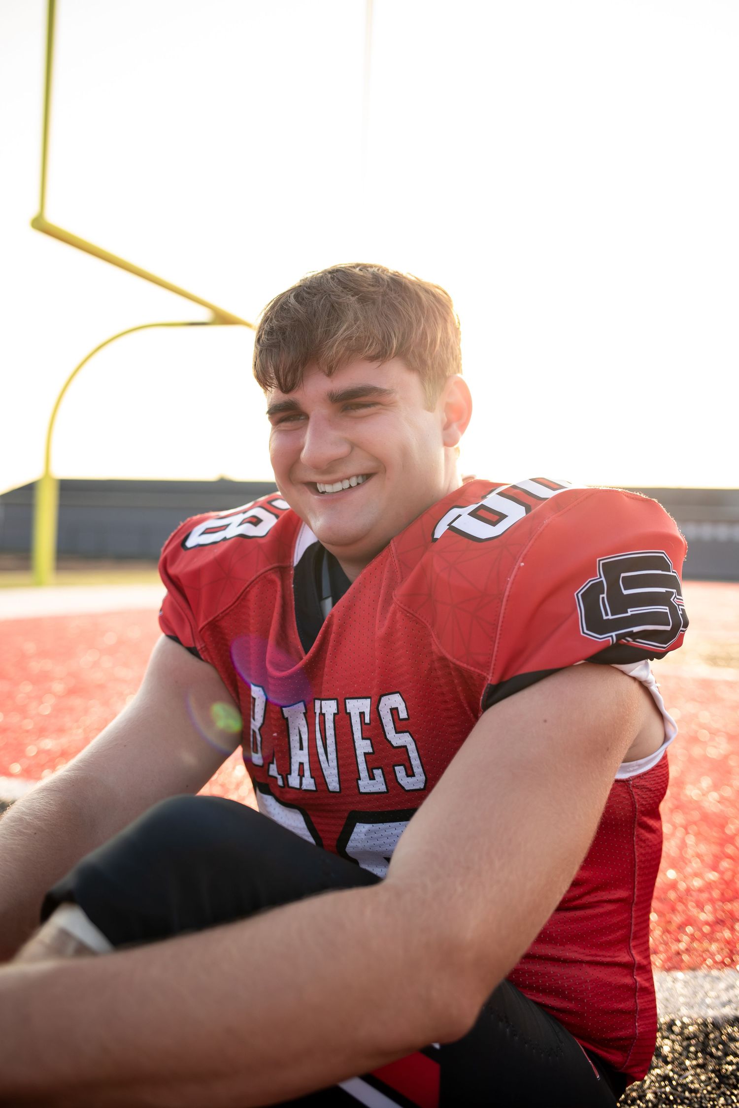 High school football player in red jersey smiling while sitting on field during practice session.