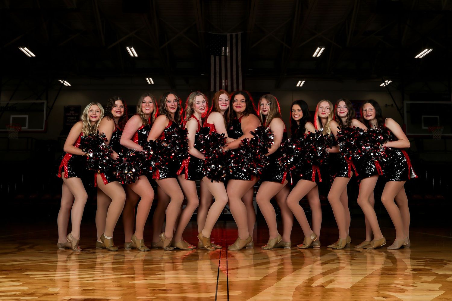 Dance team in matching black costumes poses together on wooden gymnasium floor during team photo session.
