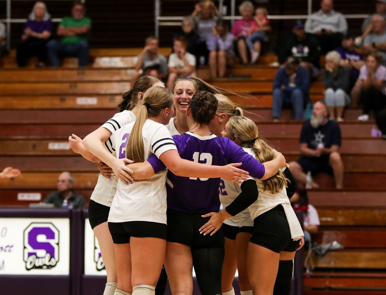 High school volleyball players huddle together in celebration during a match in a school gymnasium.