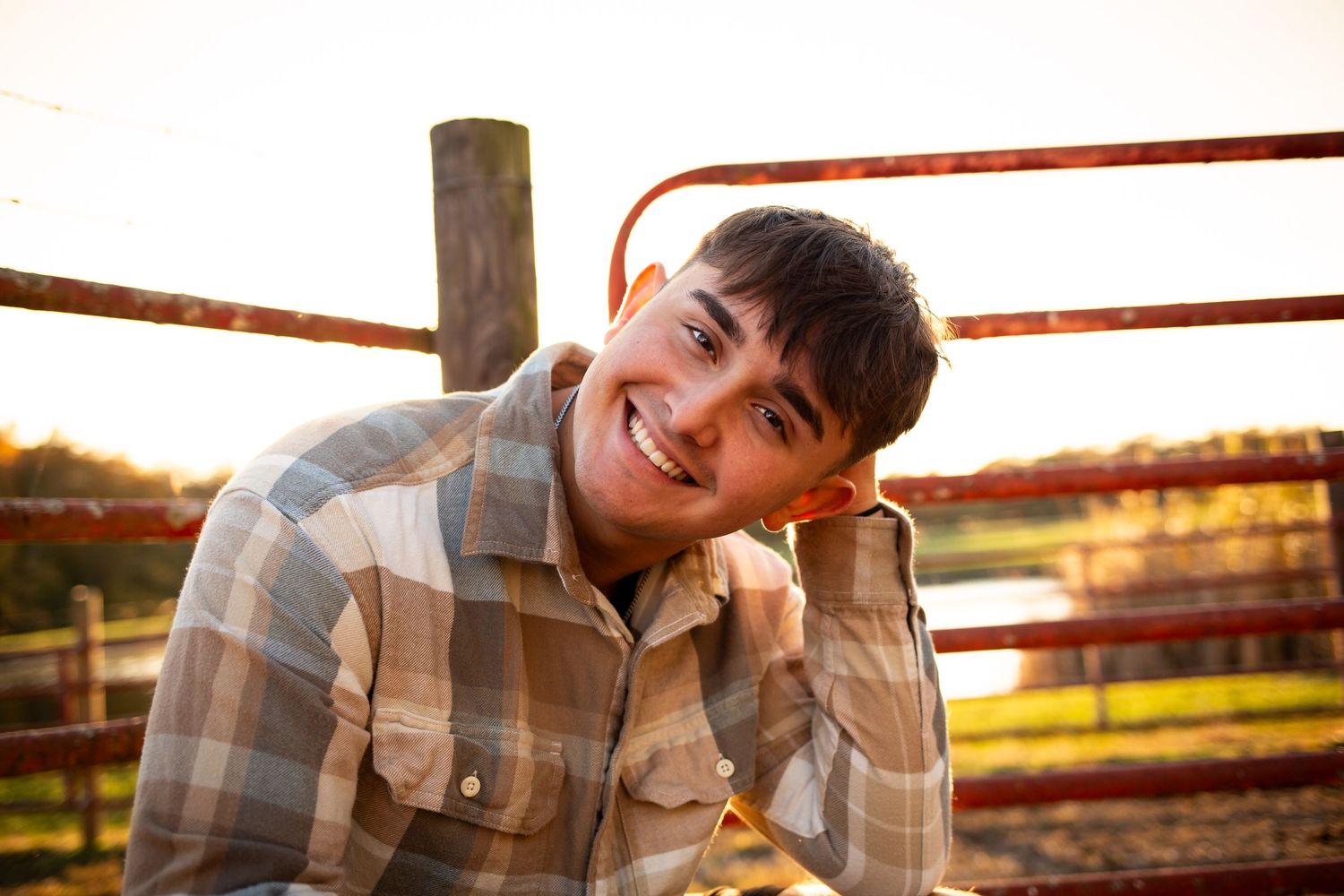 A person in a plaid shirt leans against a wooden fence at sunset in a rural setting.