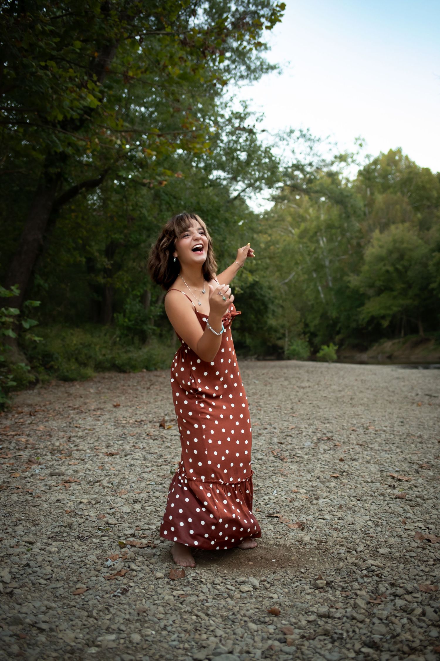 A person in a brown polka dot dress sits and laughs joyfully on a rocky ground surrounded by trees.
