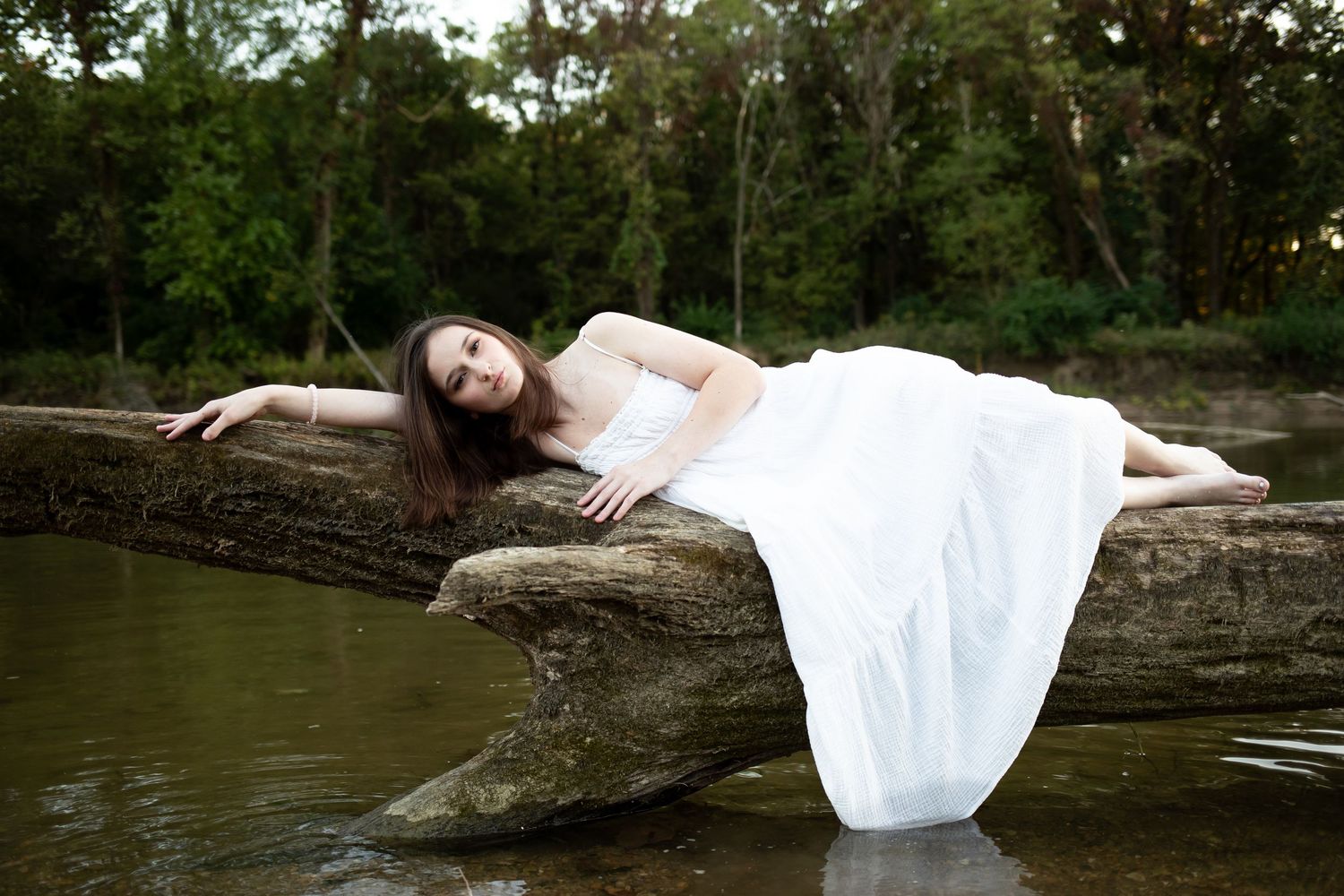 A person in a flowing white dress poses gracefully on rocks by a serene lake during a photo session.