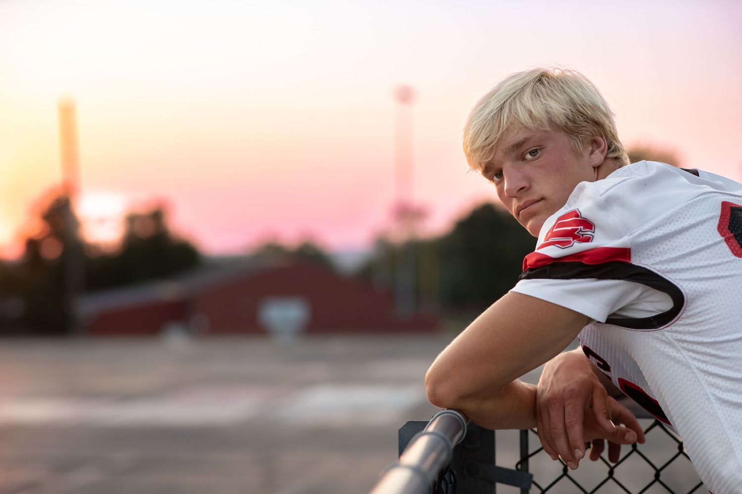 High school athlete in white football jersey leans against chain link fence at sunset during practice.
