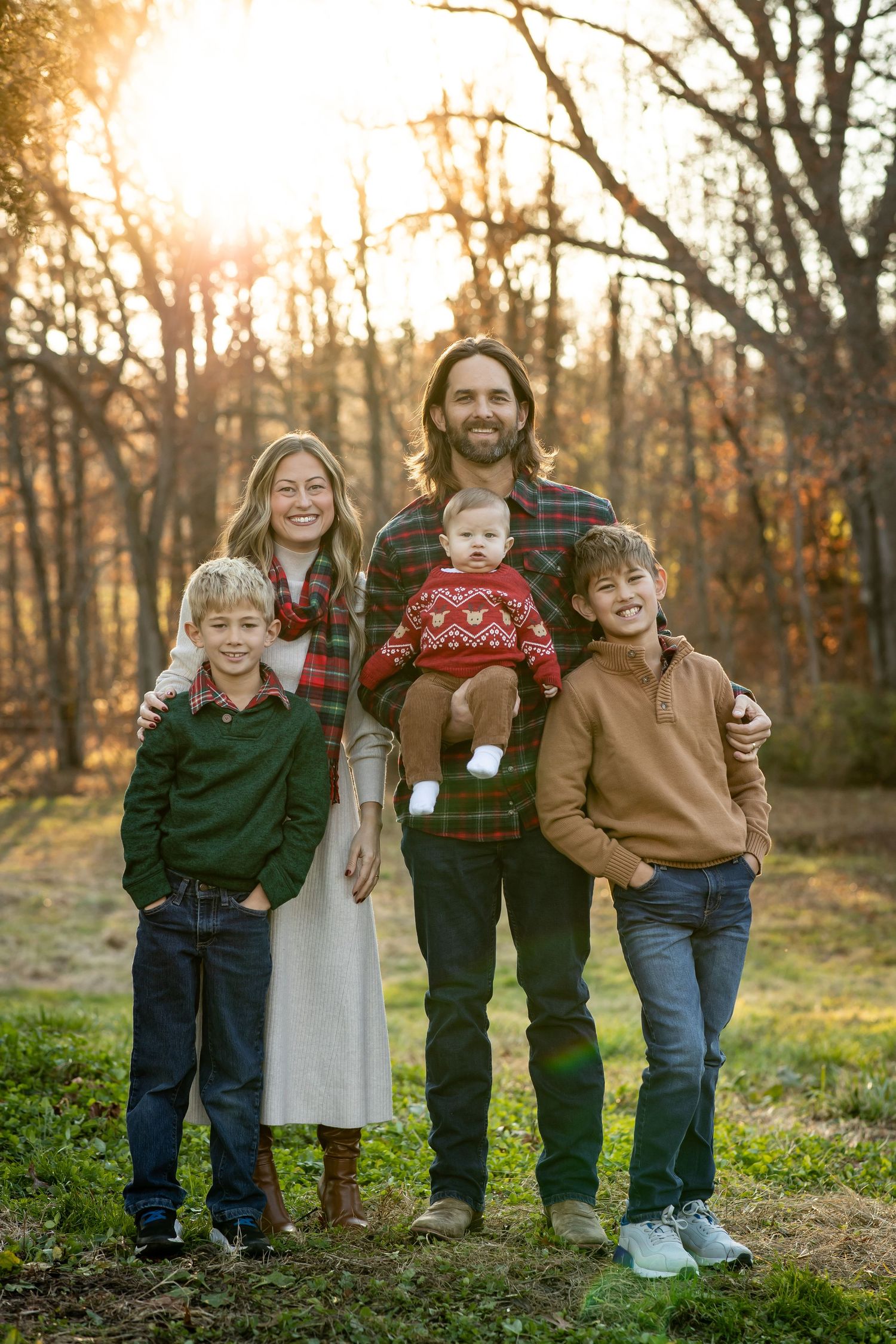A family wearing coordinated casual outfits poses together in a wooded setting during golden hour sunset.