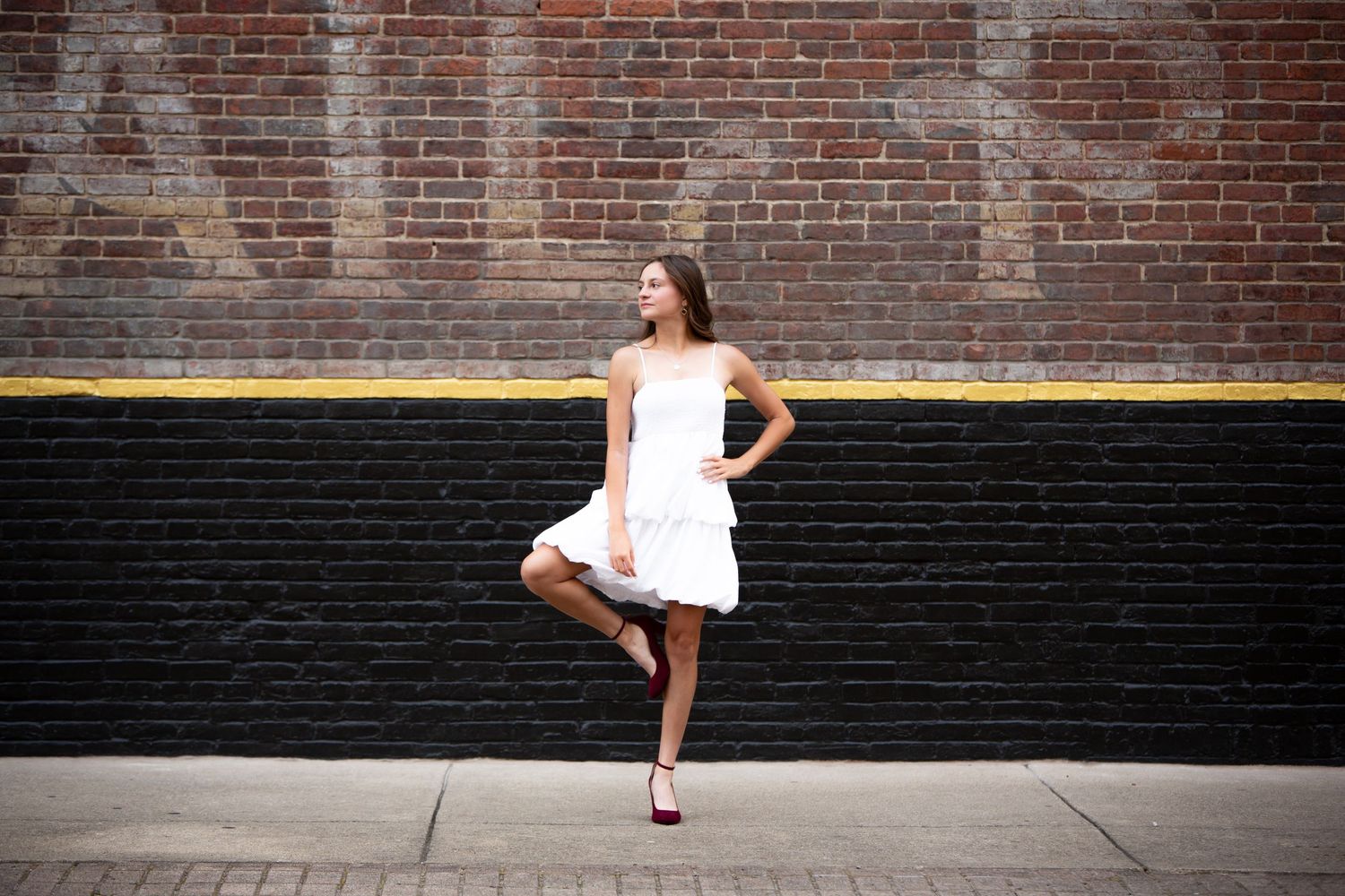 A dancer in a white dress poses gracefully against a rustic brick wall background.