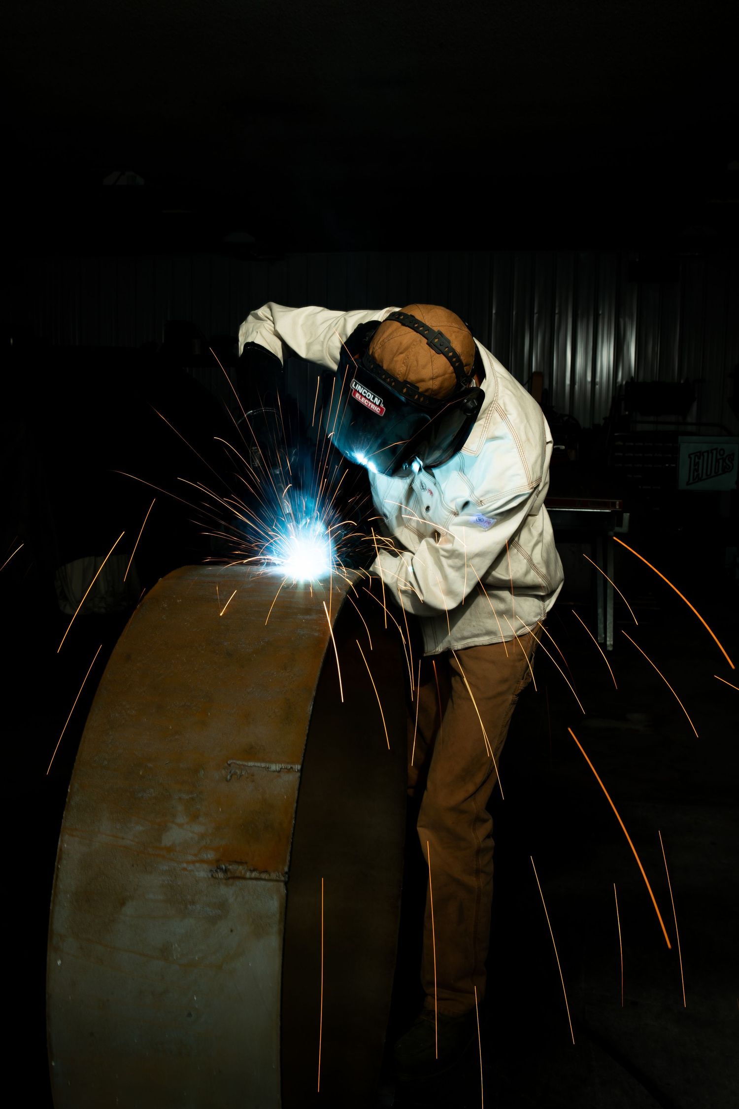 Welder in protective gear creating bright sparks while working on curved metal surfaces in dark workshop setting.