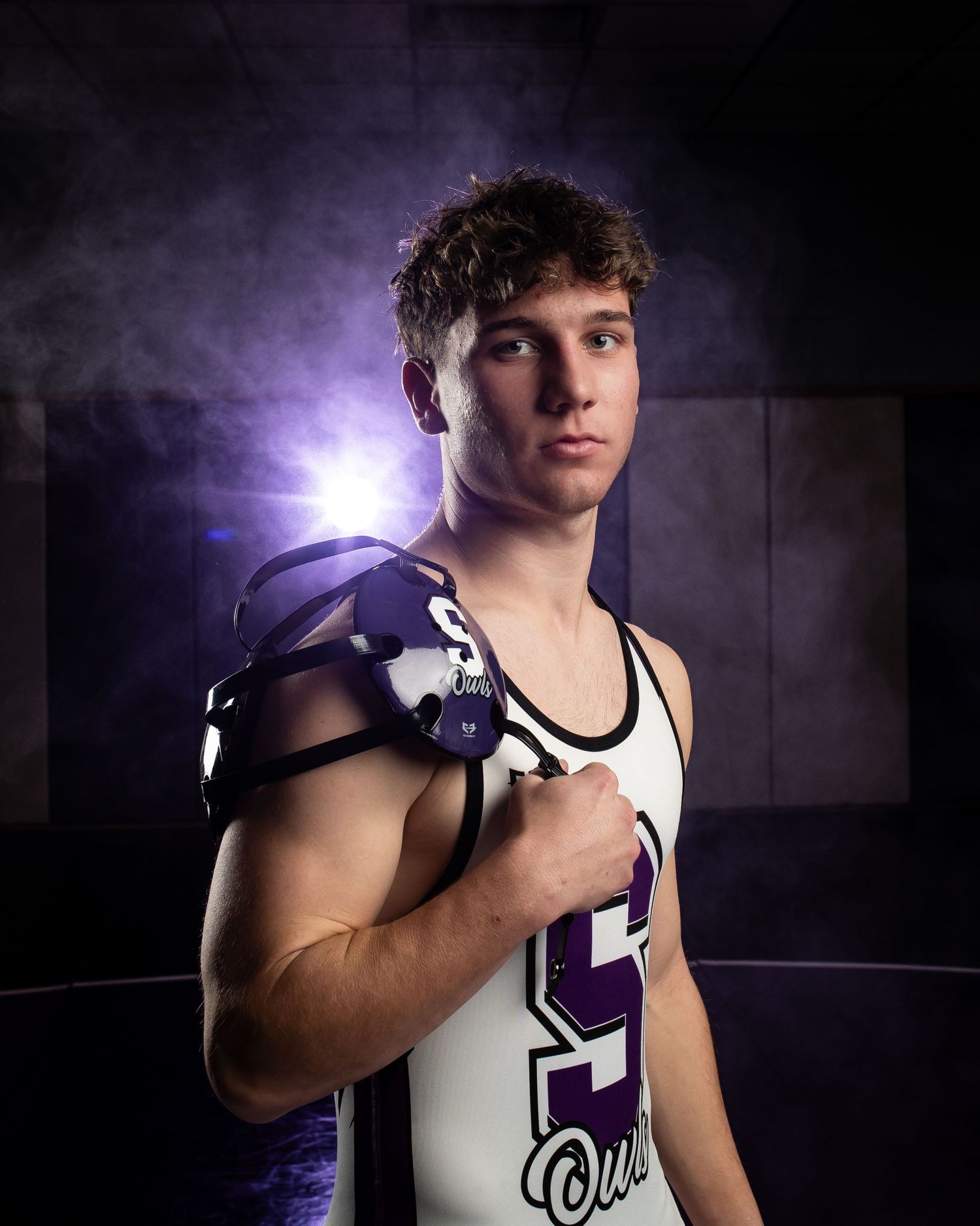 Dramatic portrait of a wrestler in purple and white uniform with moody lighting and smoke effects in the background.