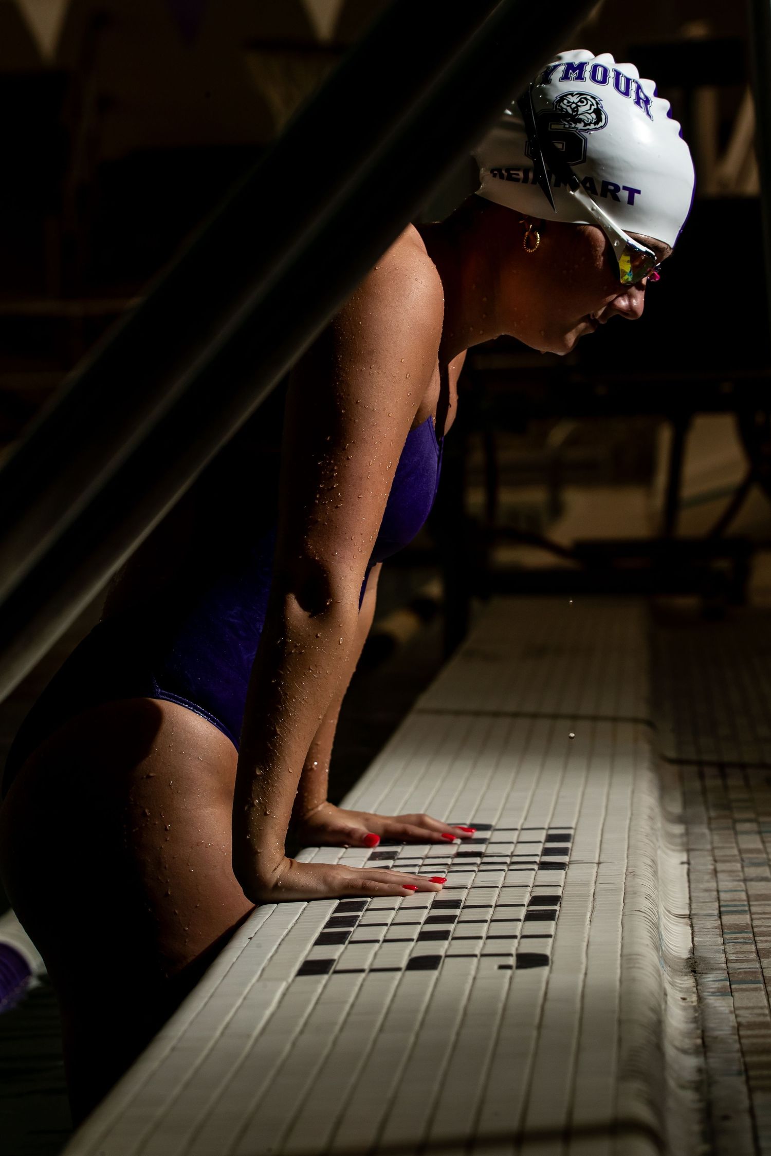 A swimmer in a white cap rests on the edge of a pool with hands on the tiled pool deck.