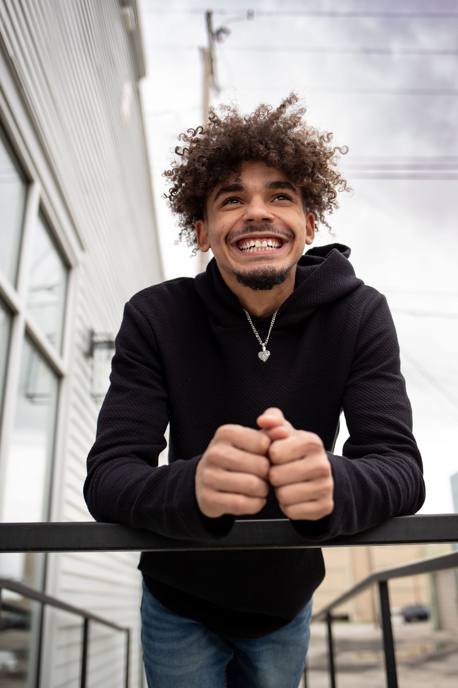 A person with curly hair wearing a black jacket leans forward while smiling and clasping their hands together.